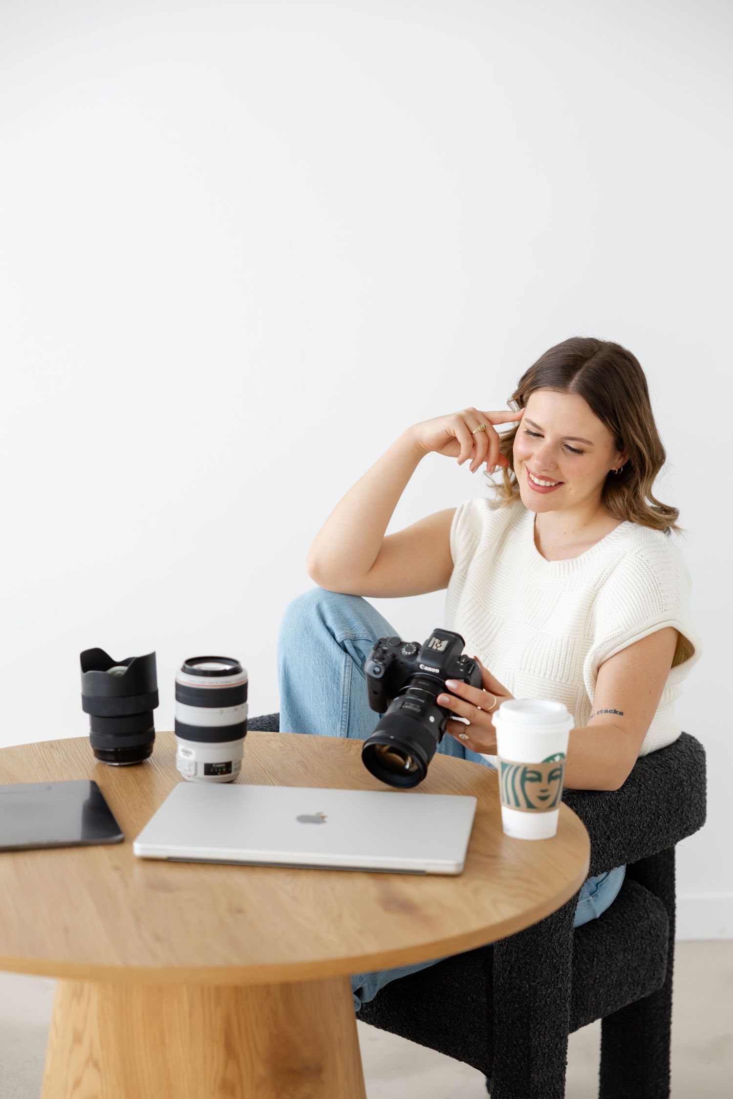 Professional photographer reviewing images on camera while working at desk with laptop and multiple lenses.