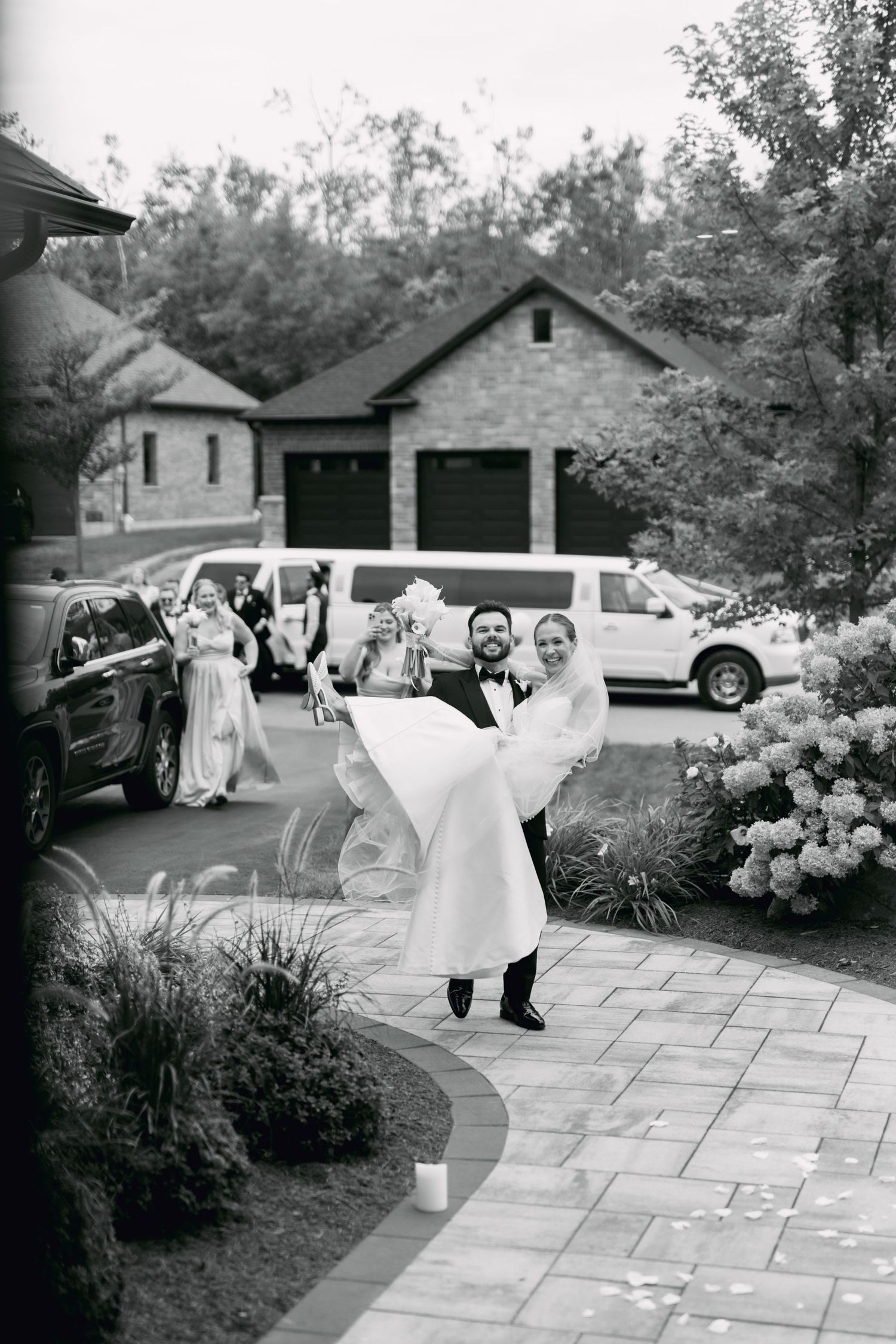 Groom carries bride in black and white photo outside wedding venue with bridal party.