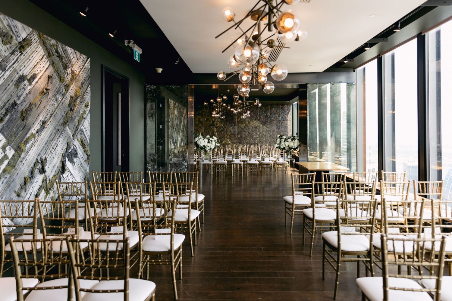 Modern wedding ceremony venue with gold Chiavari chairs, geometric chandelier, and floor-to-ceiling windows.