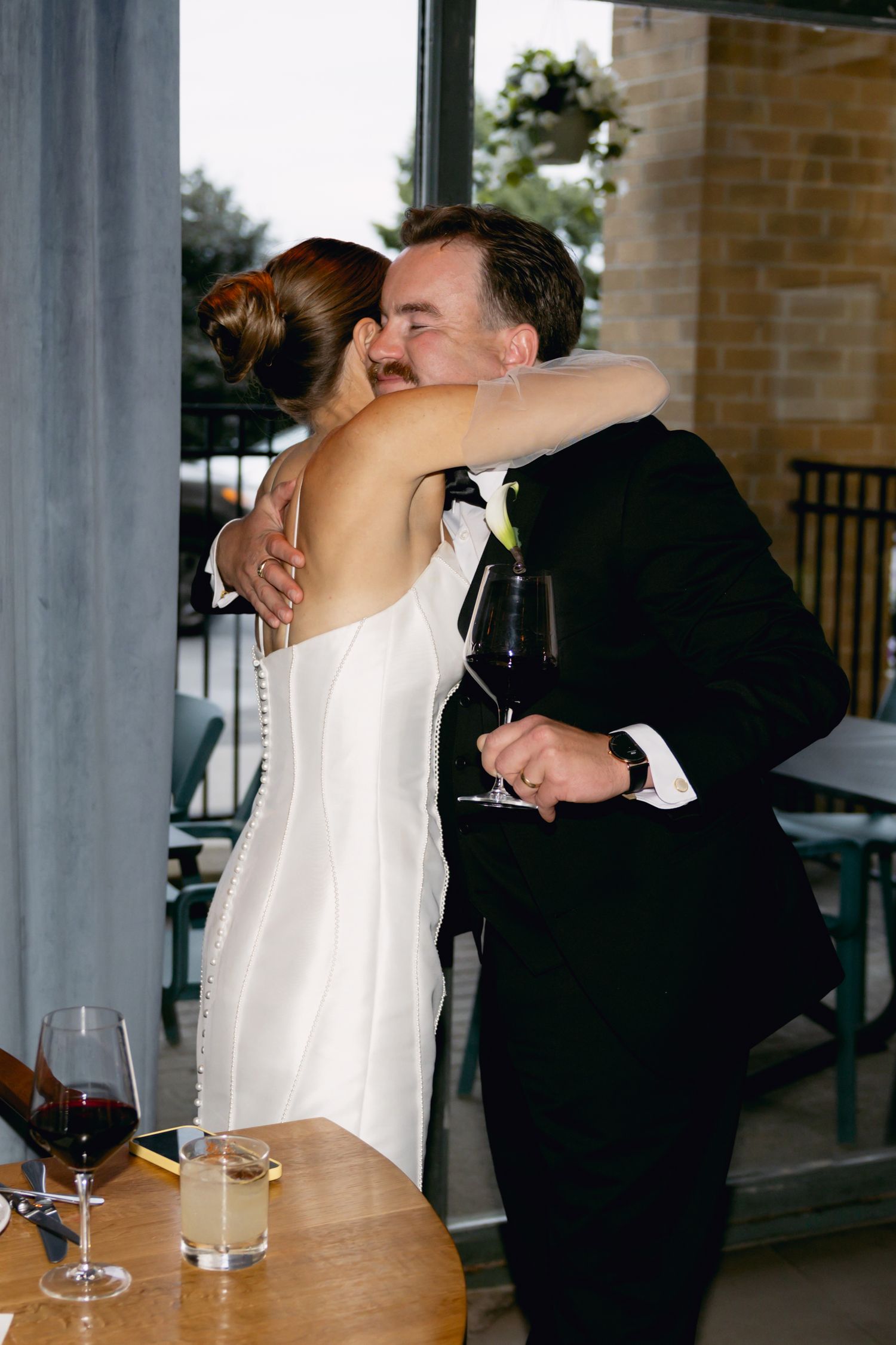 Bride in white gown and groom in black tuxedo share an emotional hug at wedding reception venue.