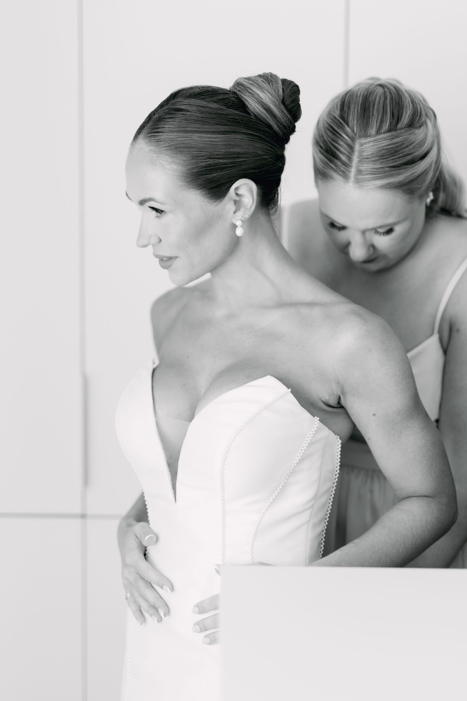 Black and white photo of bridesmaid helping bride fasten her strapless wedding gown.