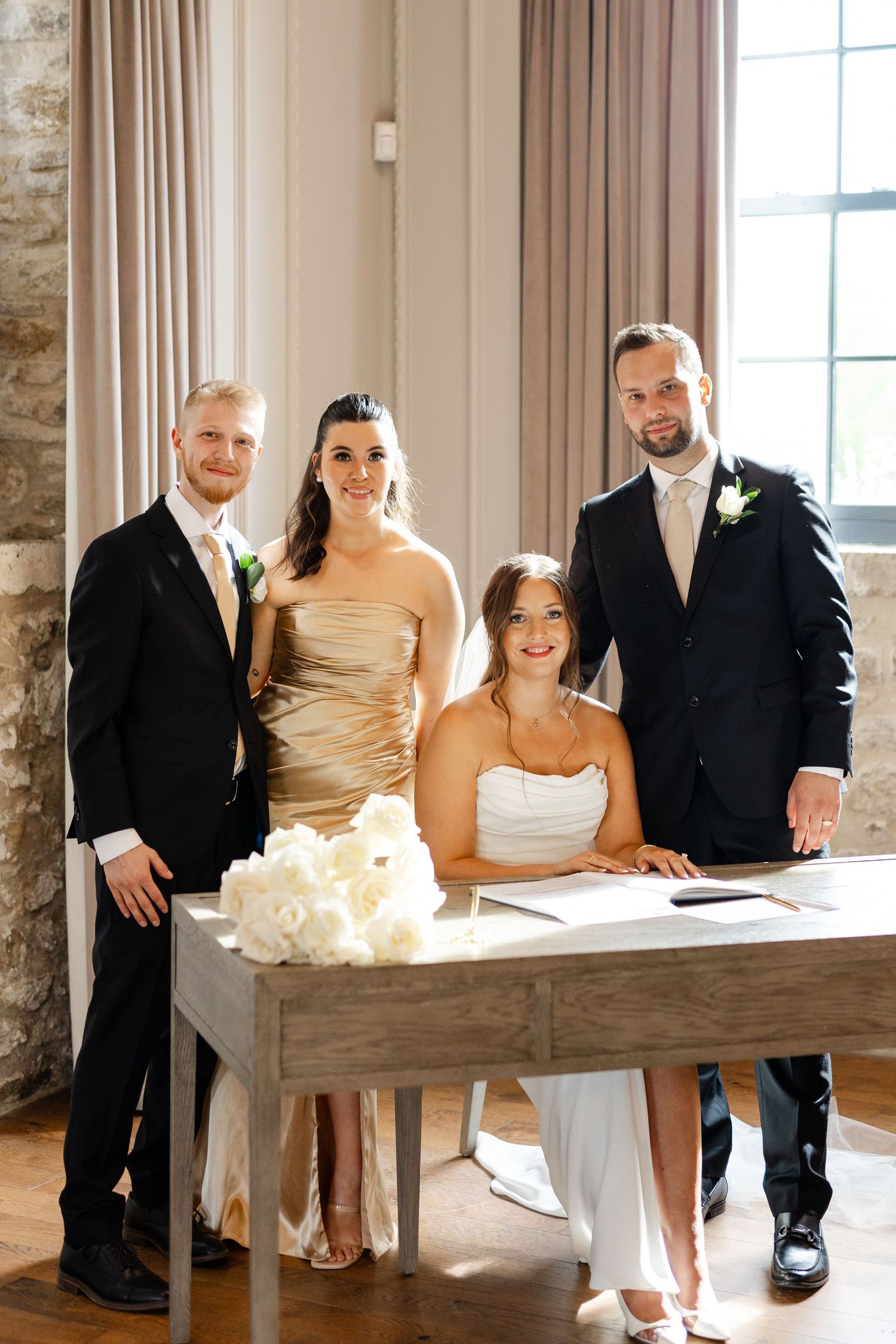 Bride and groom pose with two witnesses at marriage certificate signing table with white roses.