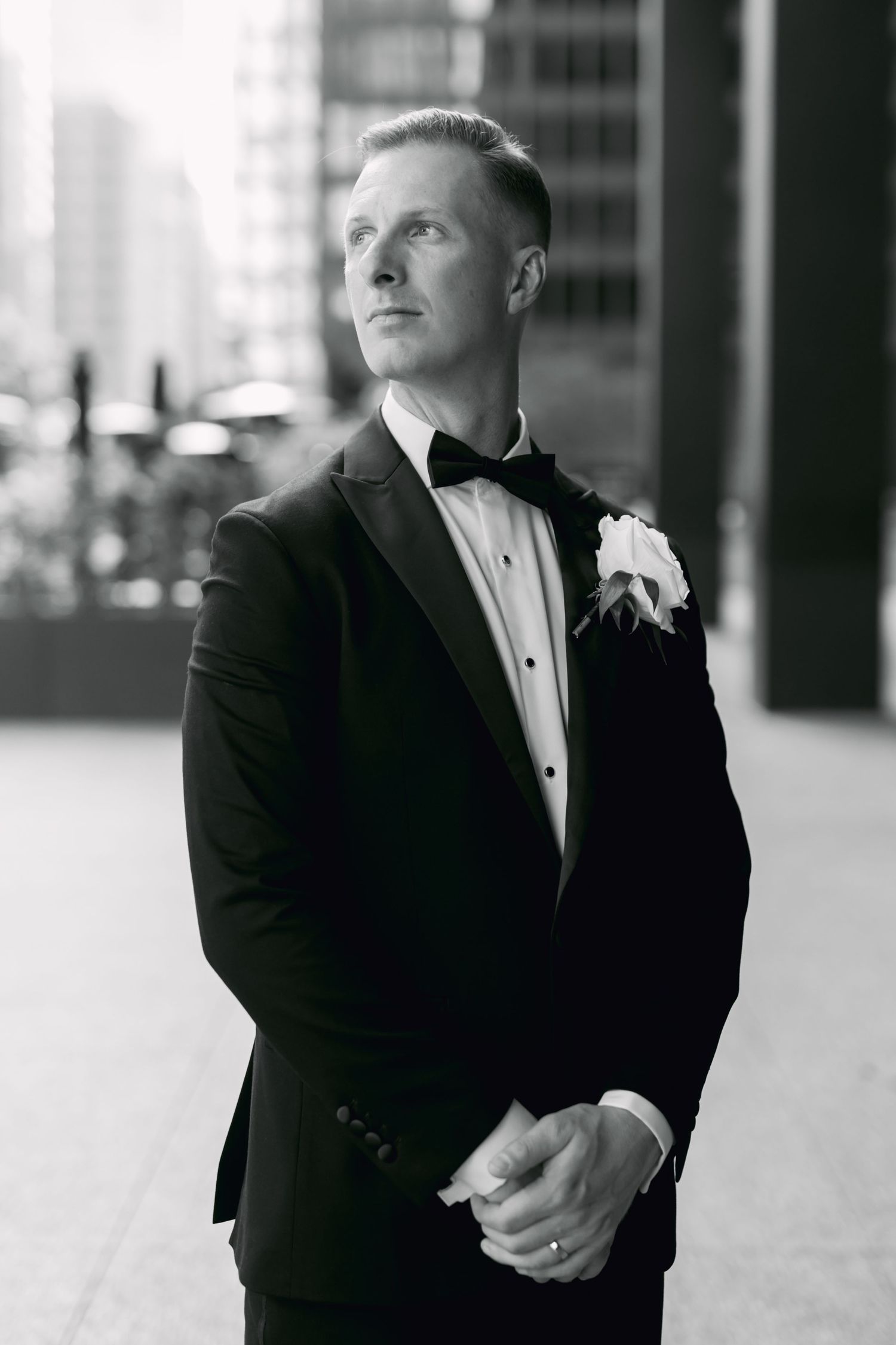 Black and white portrait of groom in tuxedo with boutonniere gazing upward on city street.