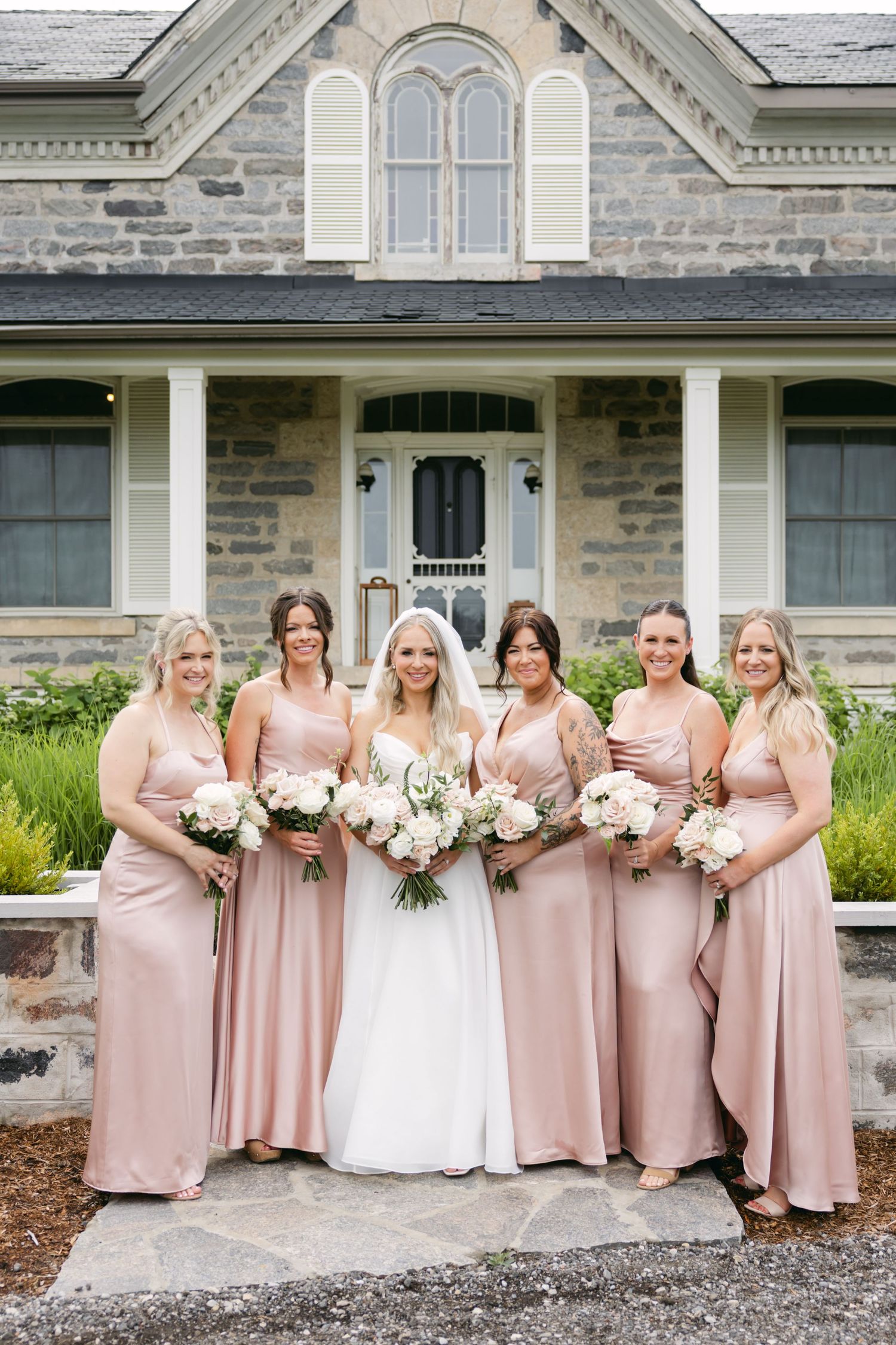 Bridesmaids in blush pink dresses pose together outside a stone building.