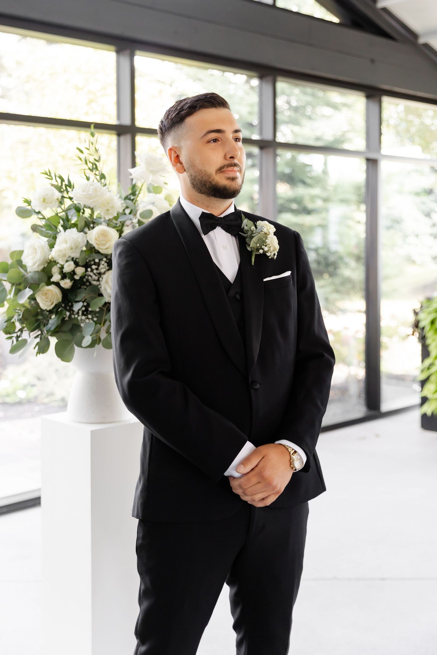 Groom in black tuxedo standing in modern white venue with floral arrangements.