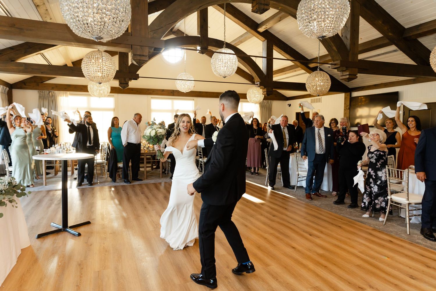 Wedding reception venue with exposed wooden beams and crystal chandeliers shows guests celebrating.