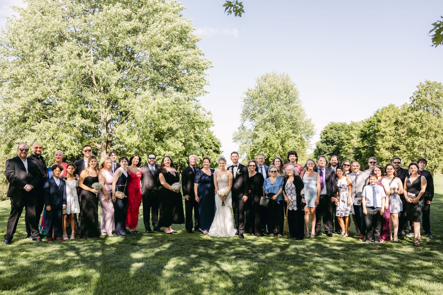 Large wedding group photo with guests arranged on grass under trees.