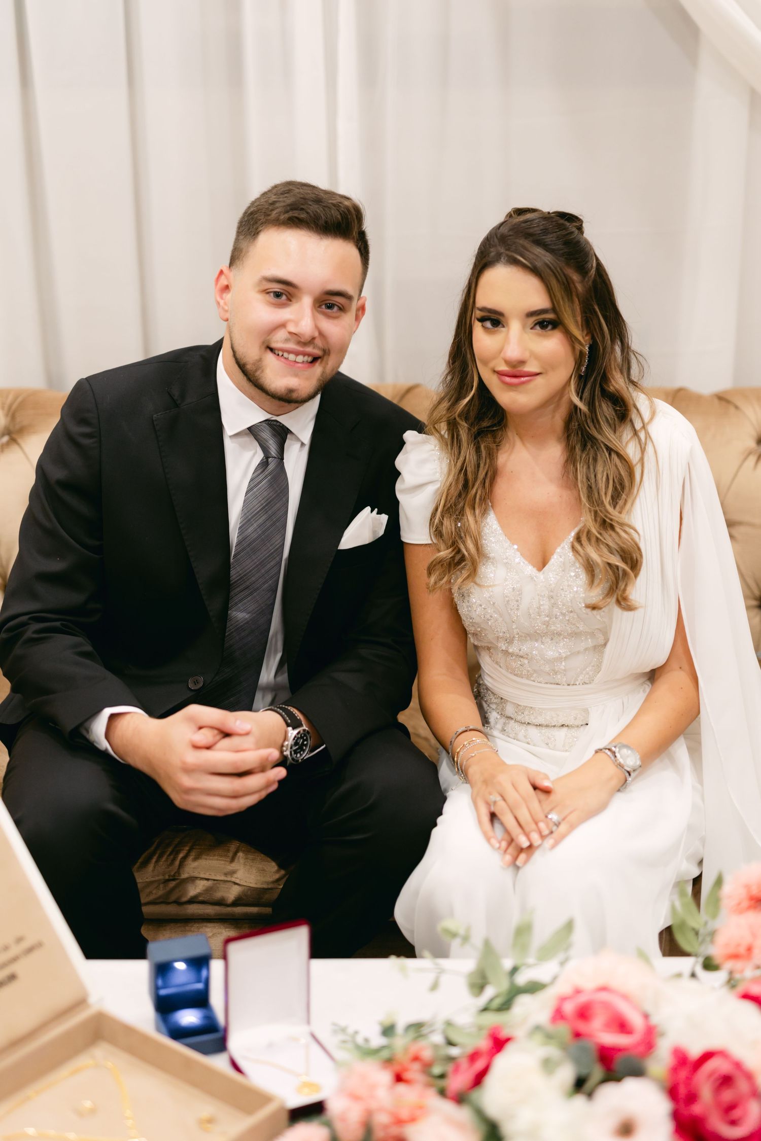 Couple sitting on beige couch during formal ceremony with floral decorations.