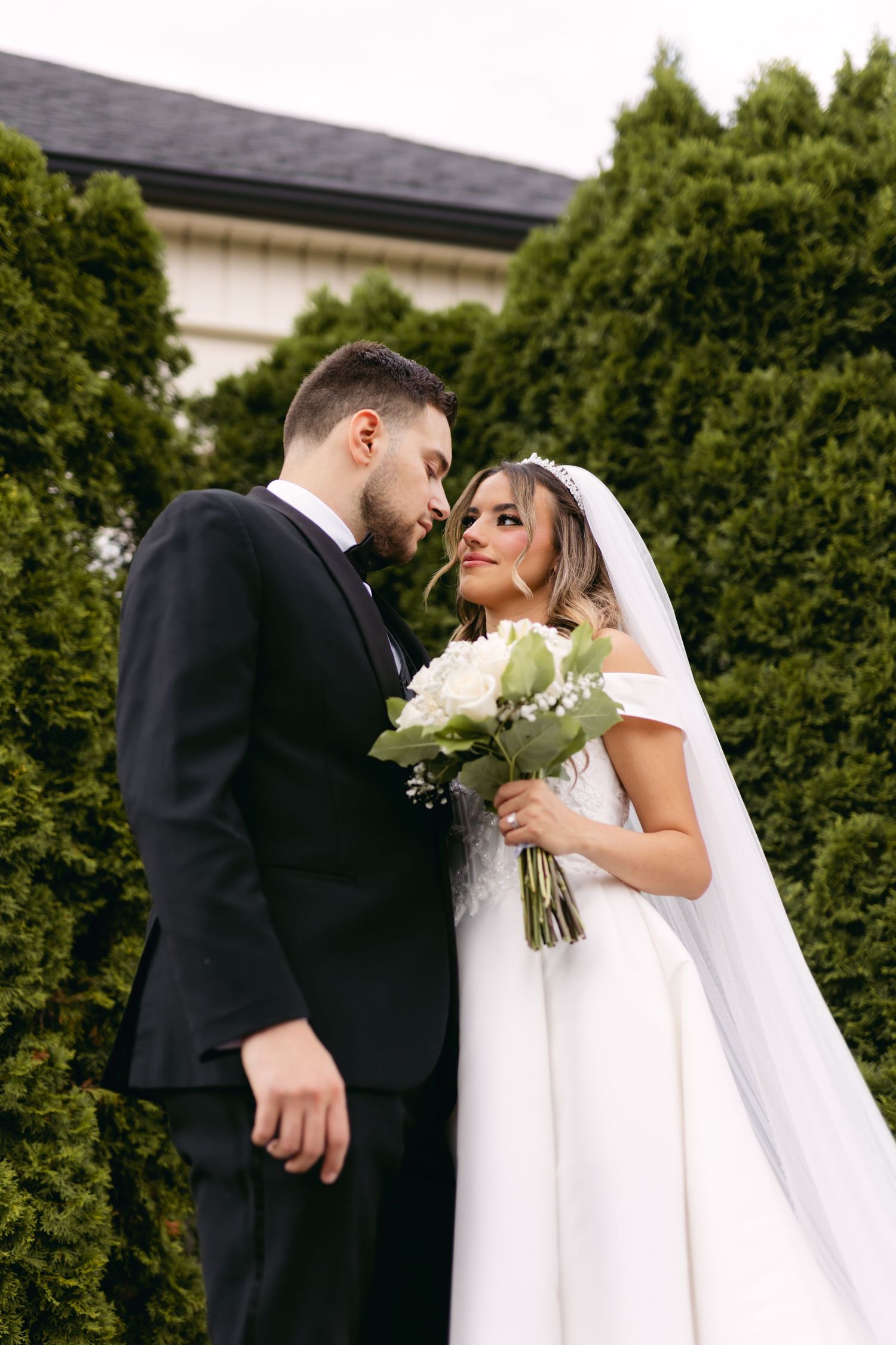 Wedding couple poses by evergreen hedge with white bouquet and flowing veil.
