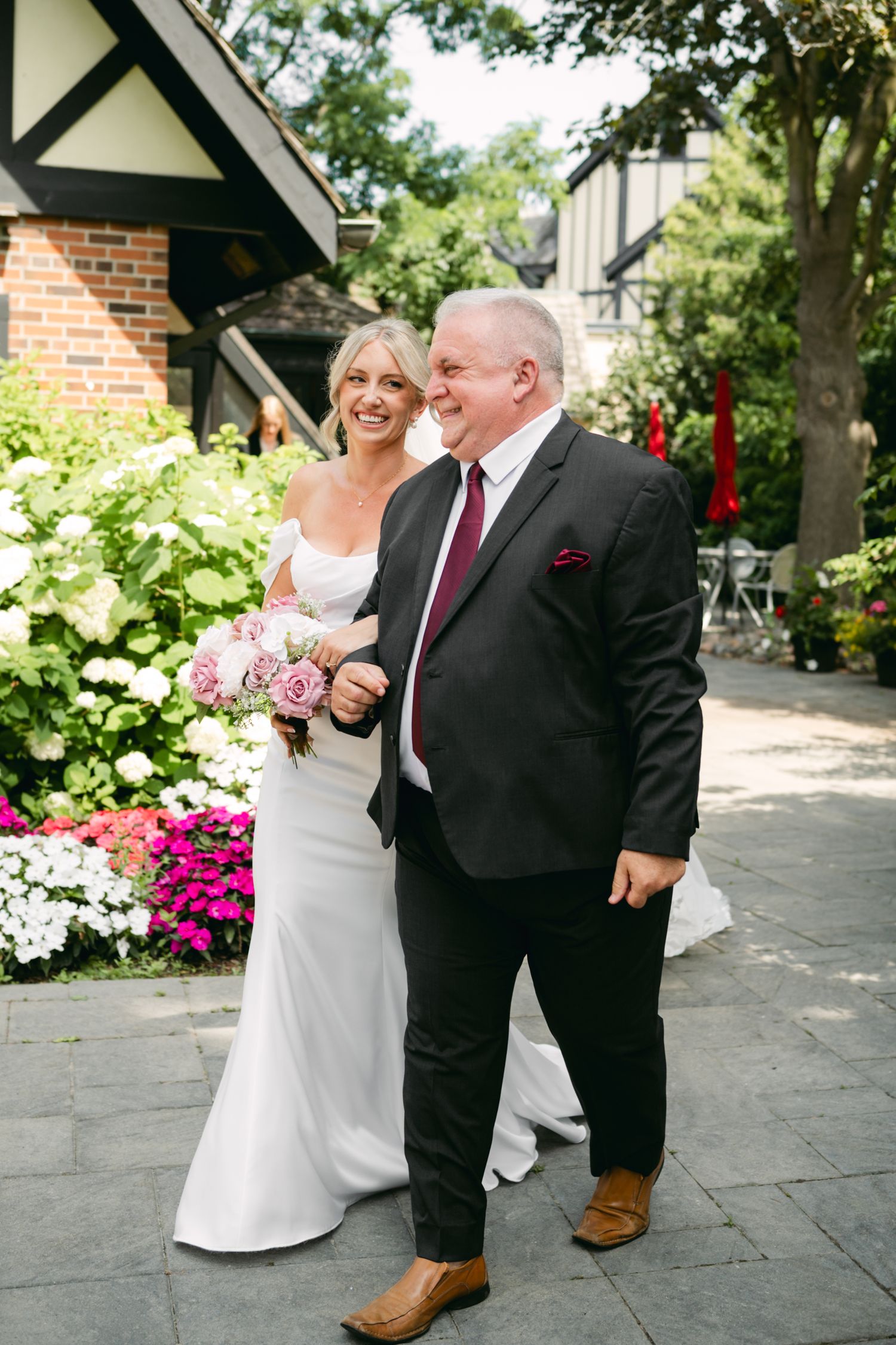 A joyful bride in a white dress walks arm-in-arm with a gentleman in a black suit at an outdoor wedding venue.