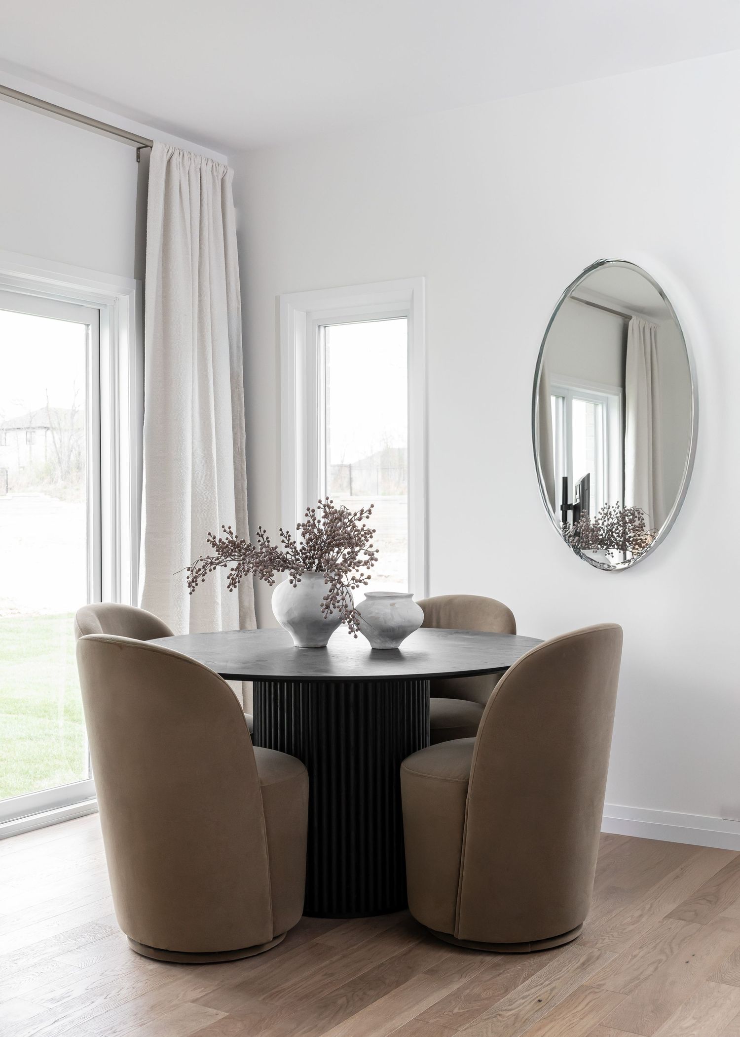Elegant dining area with round black table, beige swivel chairs, and oval mirror on white walls.