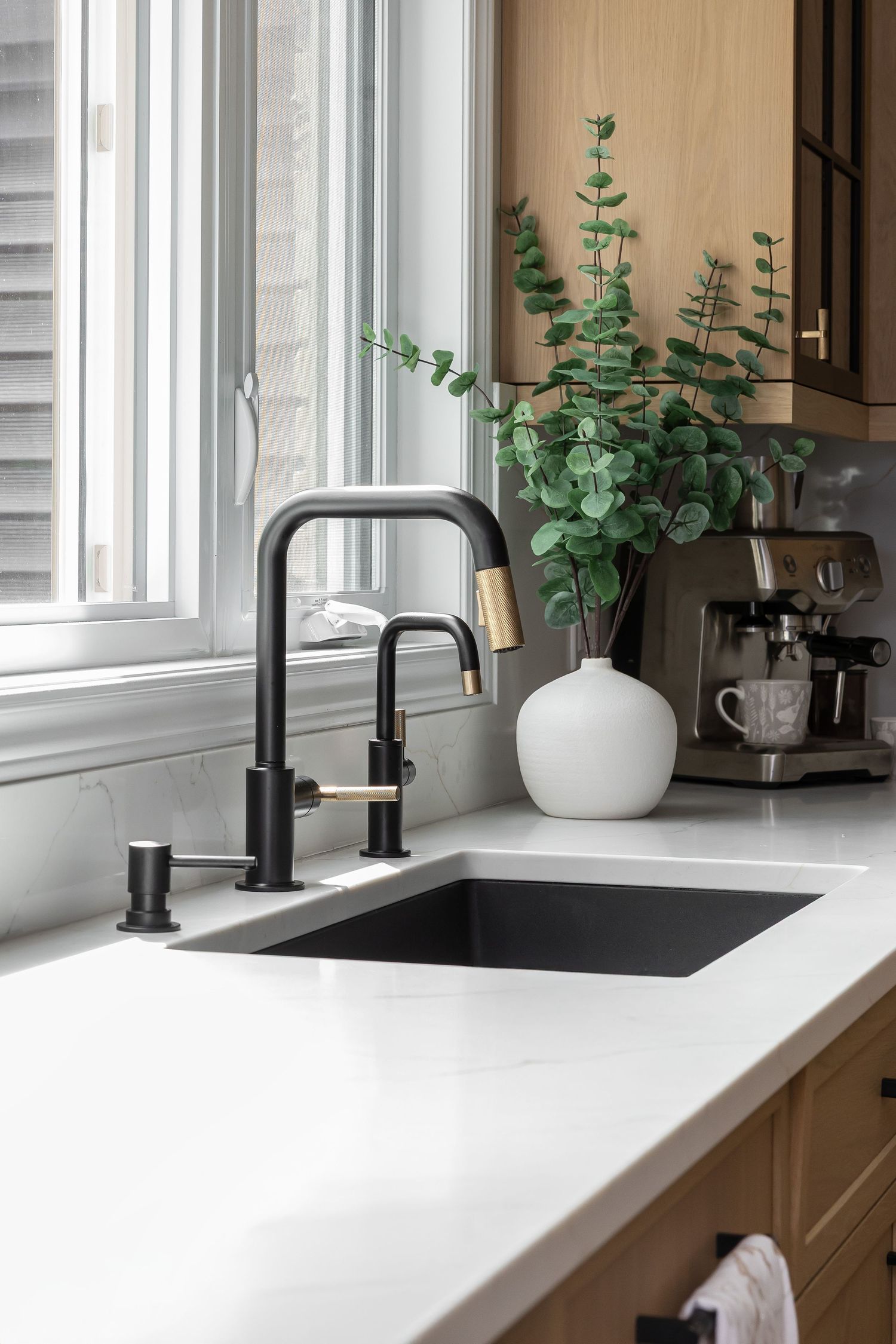 White kitchen sink with matte black faucet and eucalyptus plant decor by window.