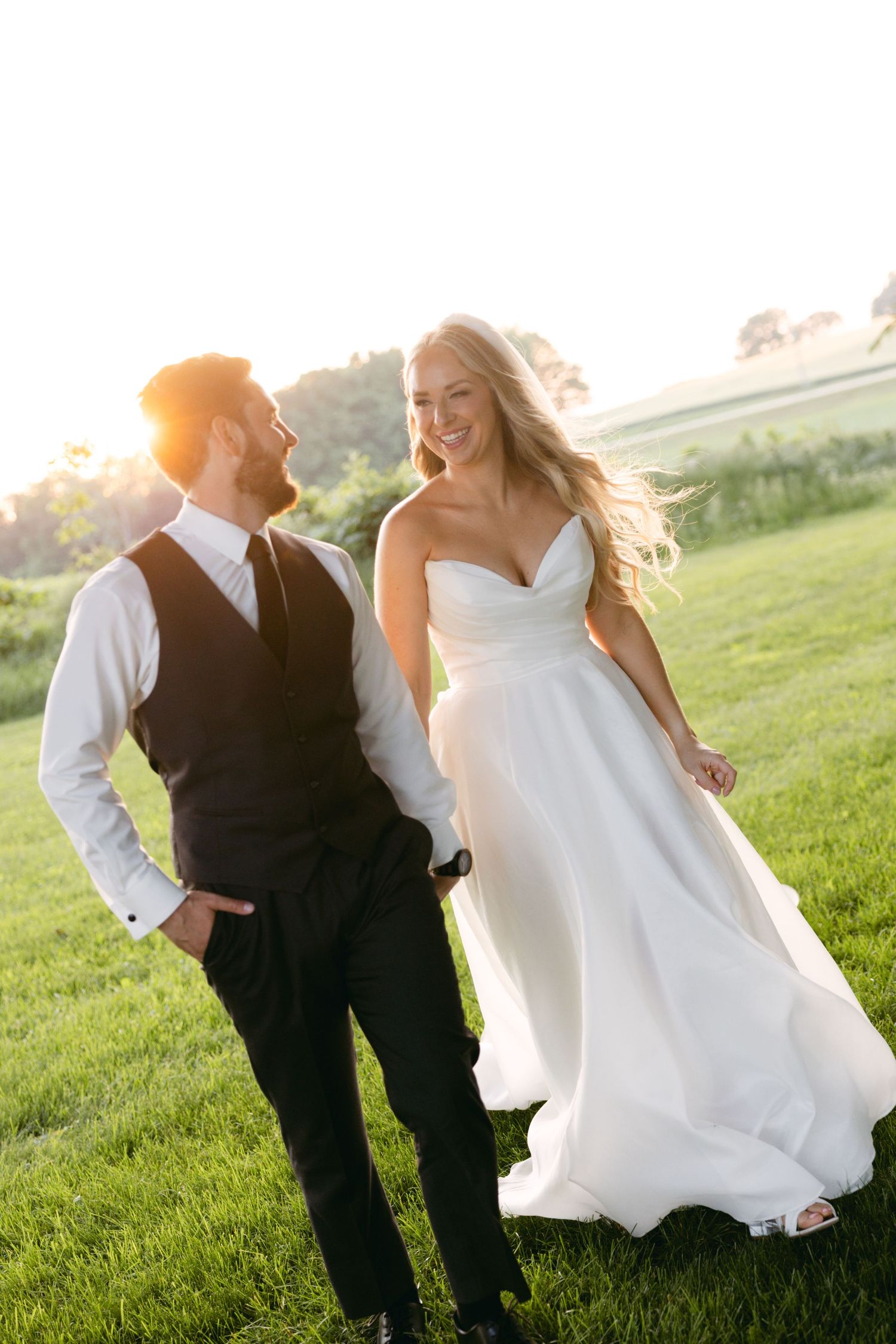 Playful wedding couple runs through grass field during golden hour photoshoot.