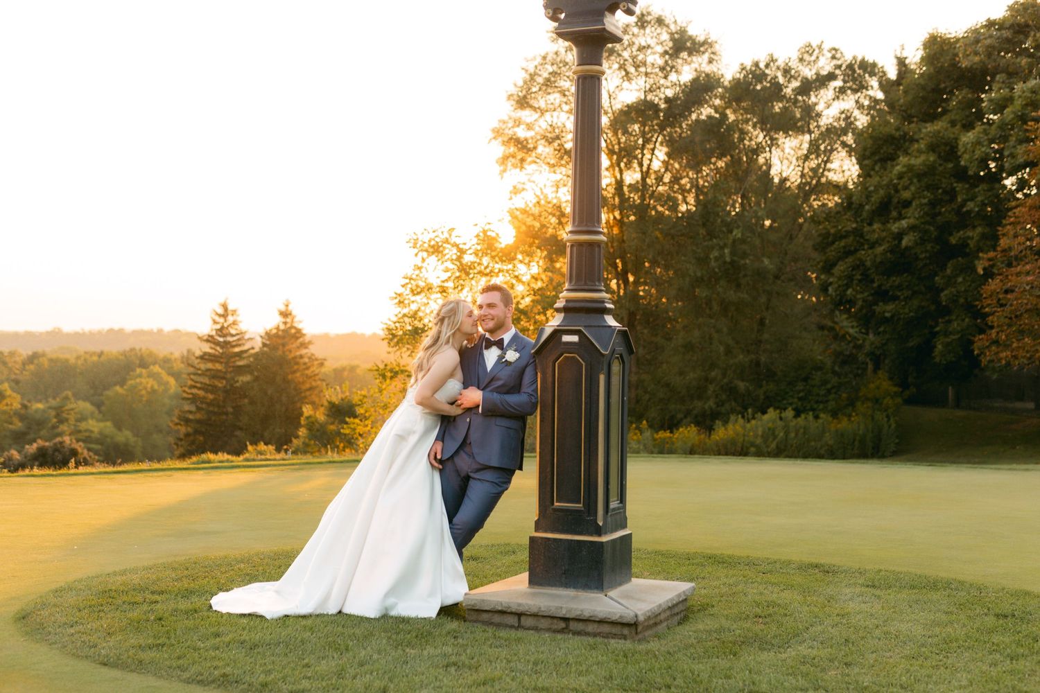 Newlyweds pose by vintage lamppost on golf course during golden hour sunset.