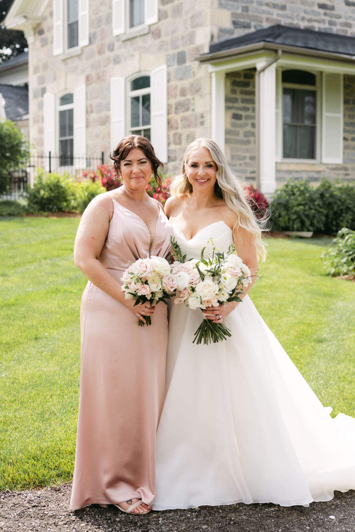 Wedding party members in blush pink and white dresses pose together on a lawn in front of a stone building.