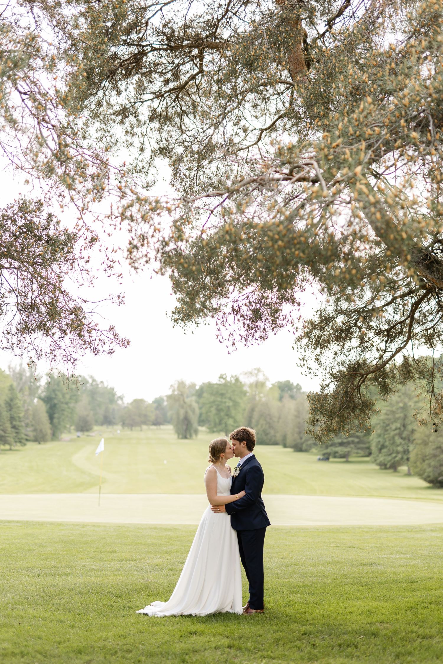 A romantic wedding couple shares an intimate moment under a tree in a scenic green field.