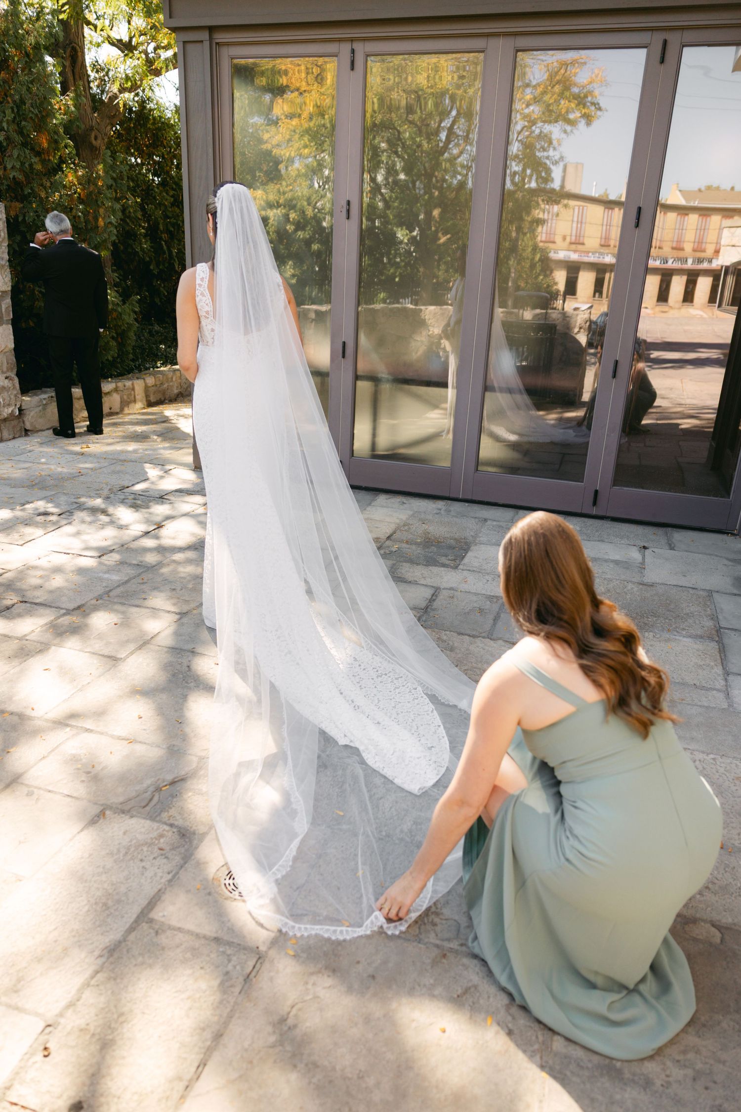 A bridesmaid in sage green adjusts a long white wedding veil outdoors.