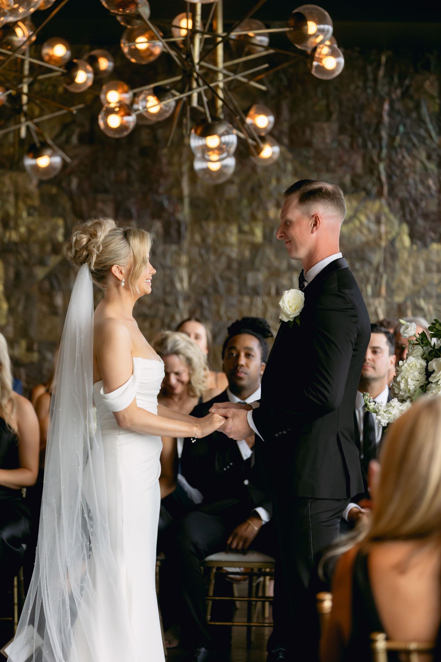 Bride and groom exchange vows at indoor wedding ceremony beneath a modern sputnik chandelier with guests seated behind.