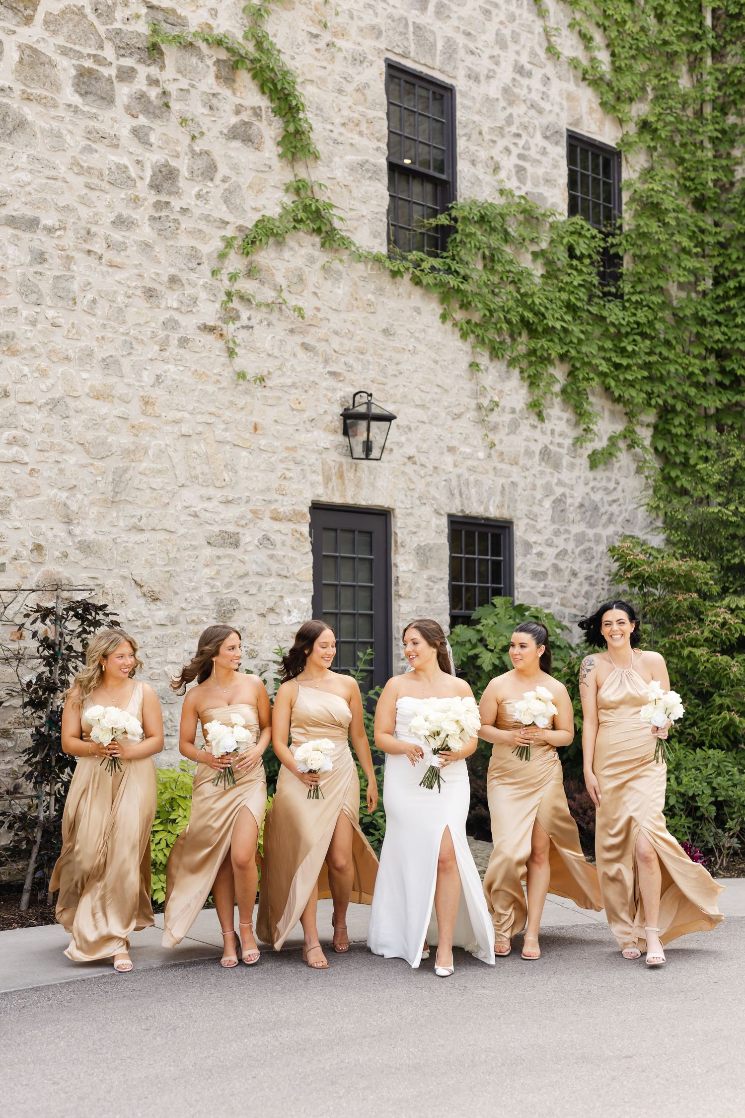 Bride in white gown walks with five bridesmaids in gold satin dresses holding white bouquets outside stone building.