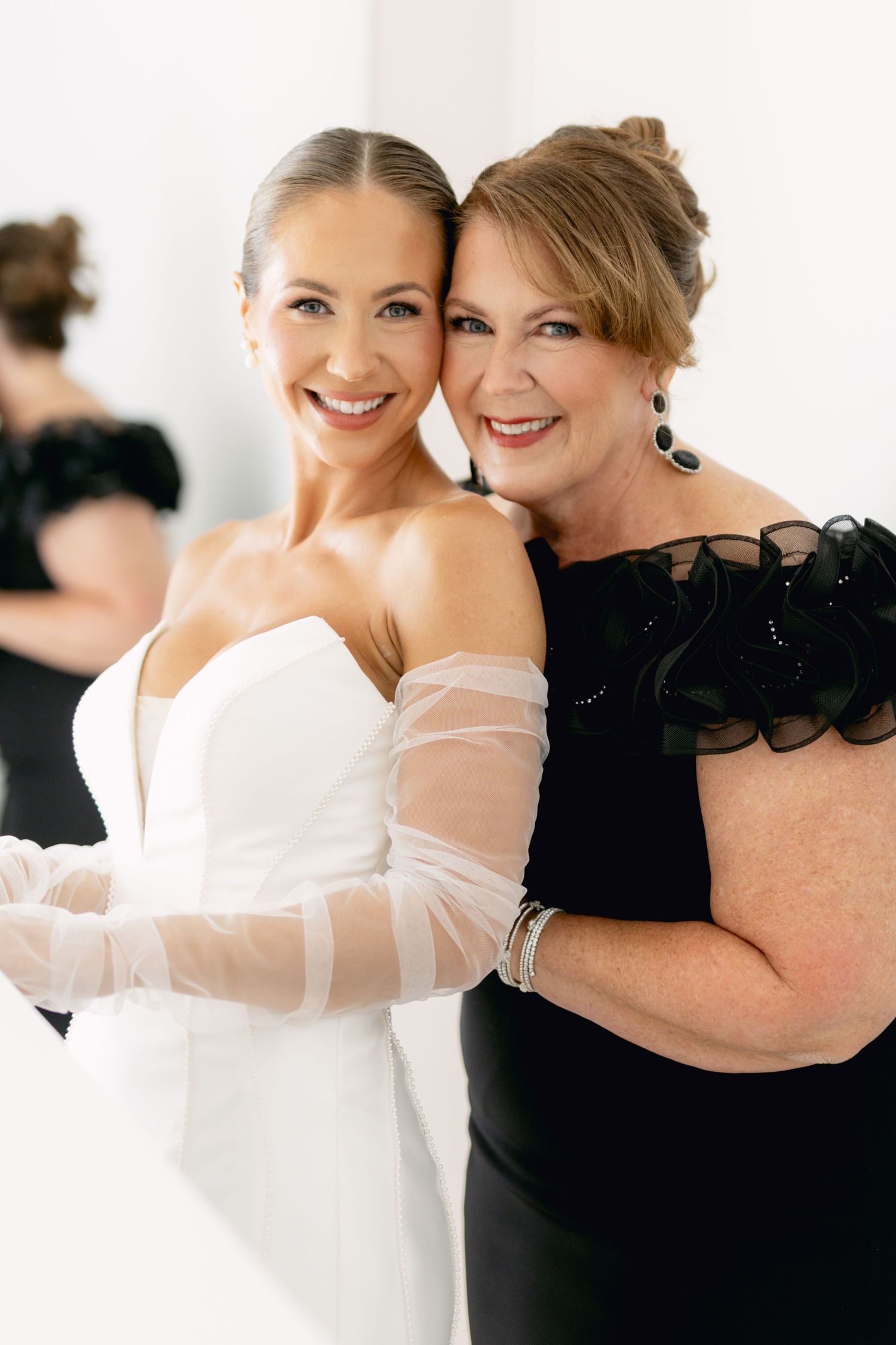 Smiling bride in white strapless gown embracing older woman in black dress during wedding preparations.