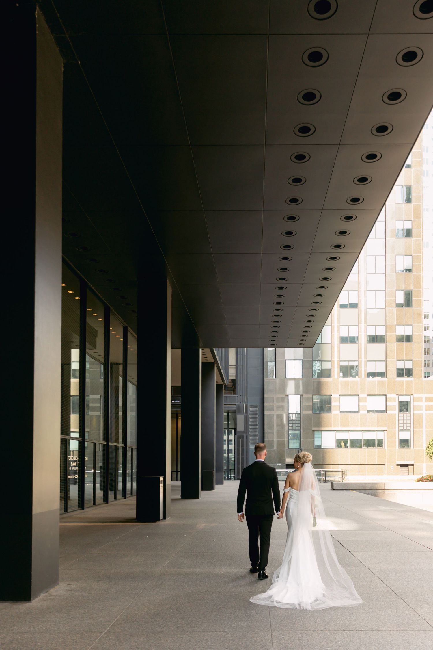 Bride and groom walk hand in hand through modern urban architectural corridor with city buildings beyond.