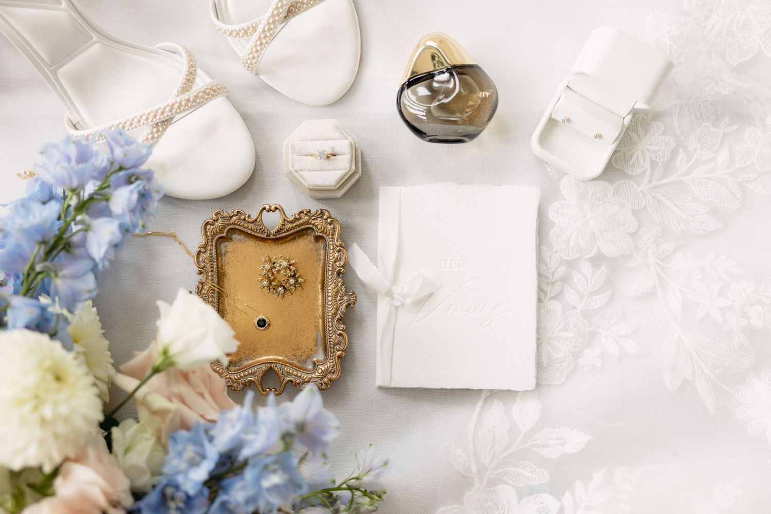 Elegant wedding details featuring delicate blue flowers, white candles, and ornate gold jewelry trays on a light surface.
