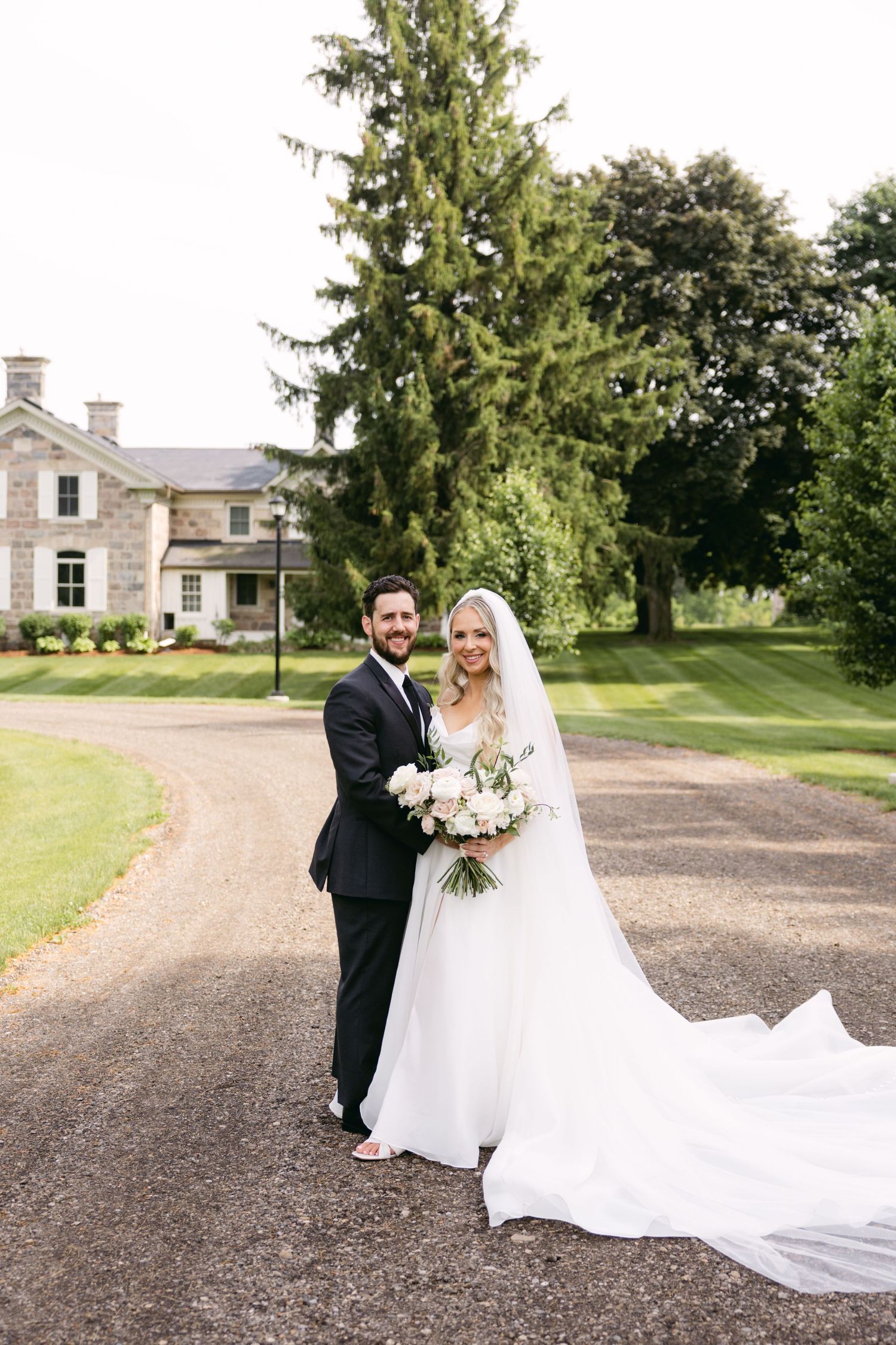 A wedding portrait on the grounds of an elegant stone estate with tall evergreen trees in the background.