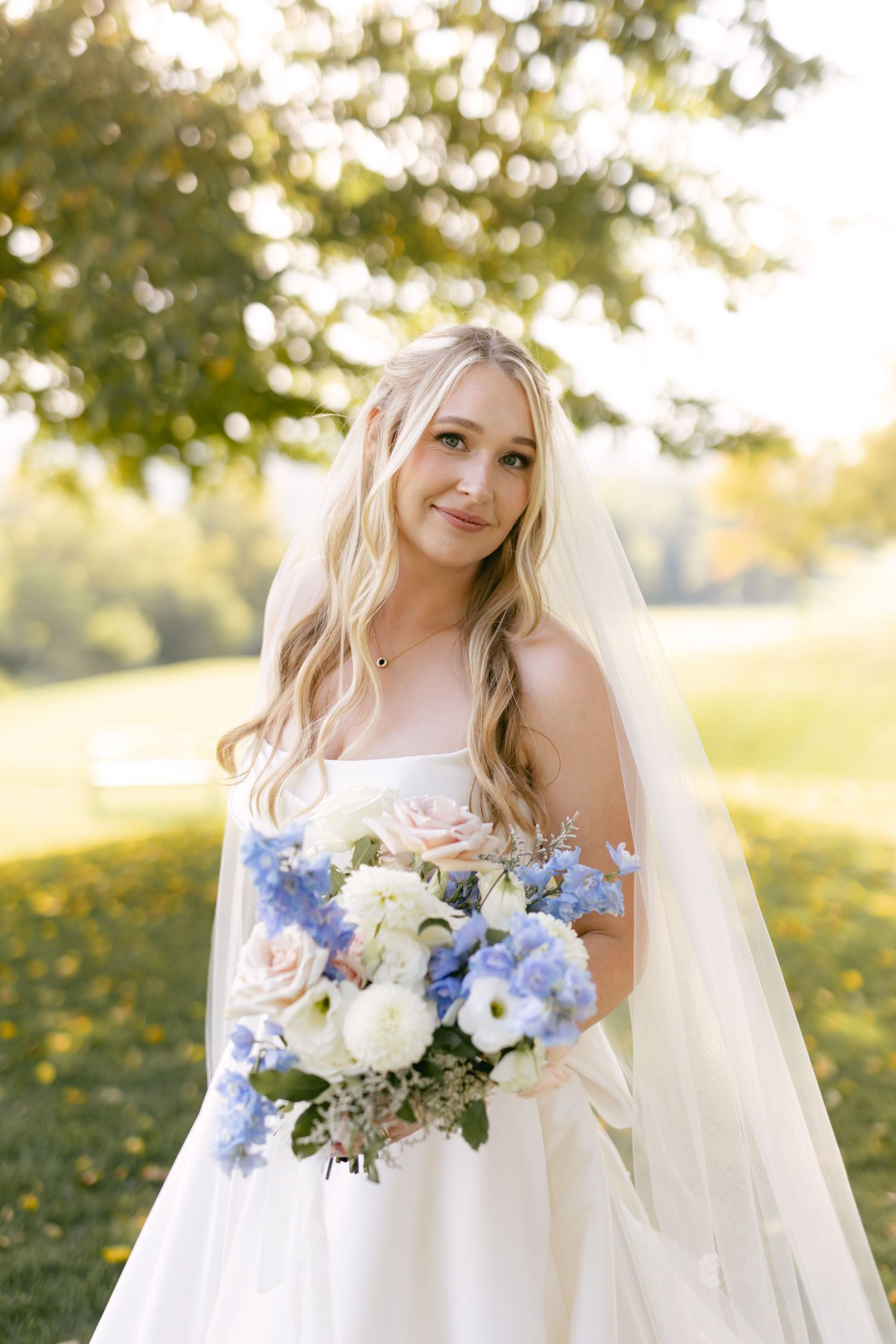 Bride with long veil holds blue and white bouquet while smiling in sunlit garden.