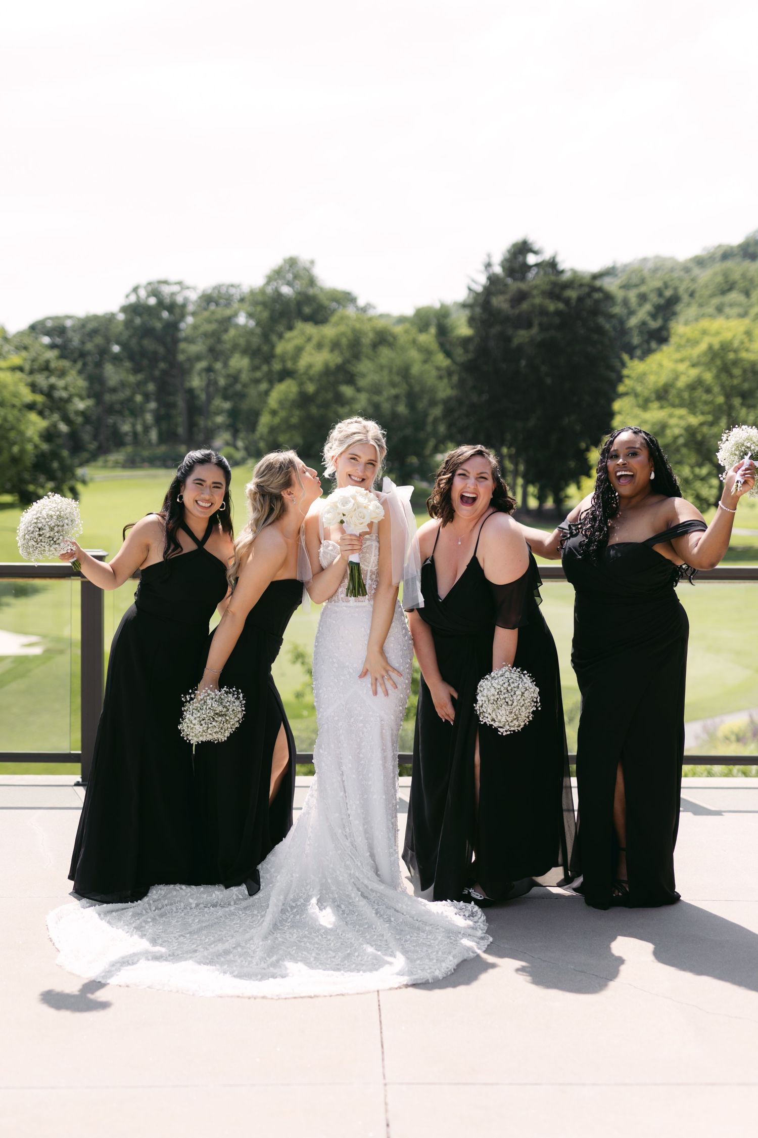 Bridal party relaxes on sunny patio wearing black dresses alongside bride in white lace gown.