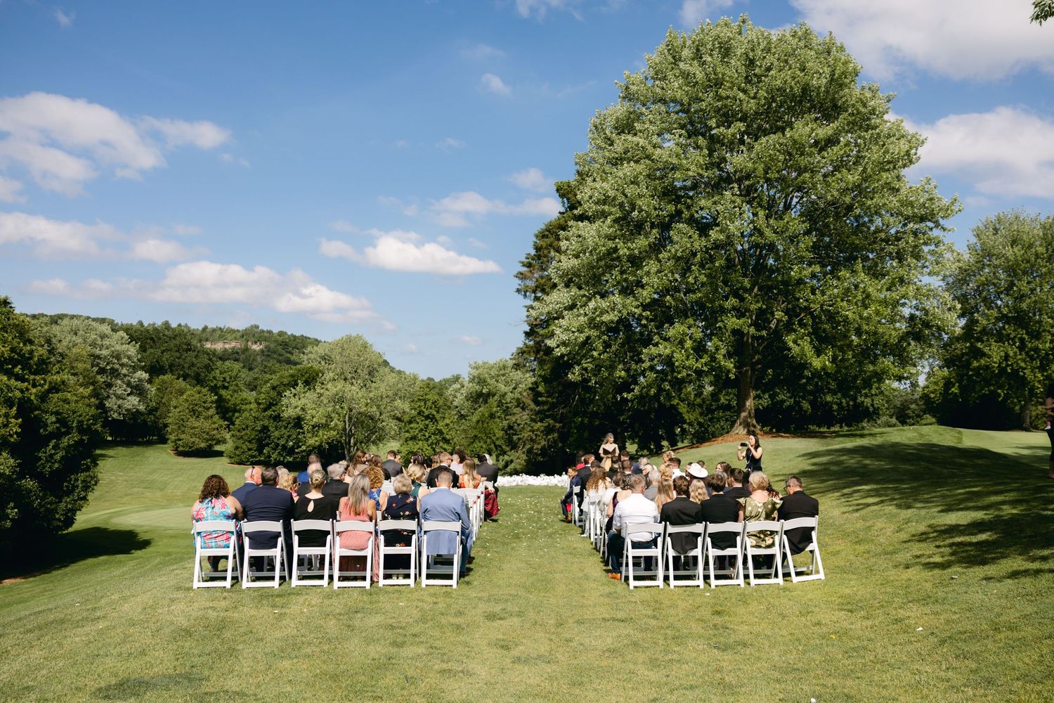Wide panoramic view of outdoor wedding ceremony setup with white chairs on a grassy hillside.