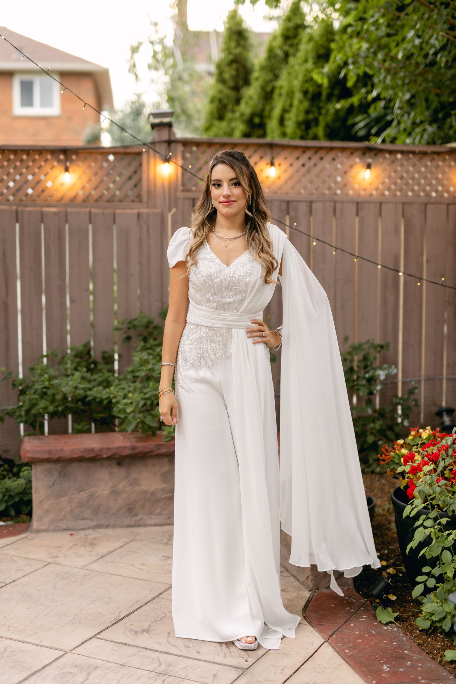Modern bride in white wedding jumpsuit with cape poses in garden setting.