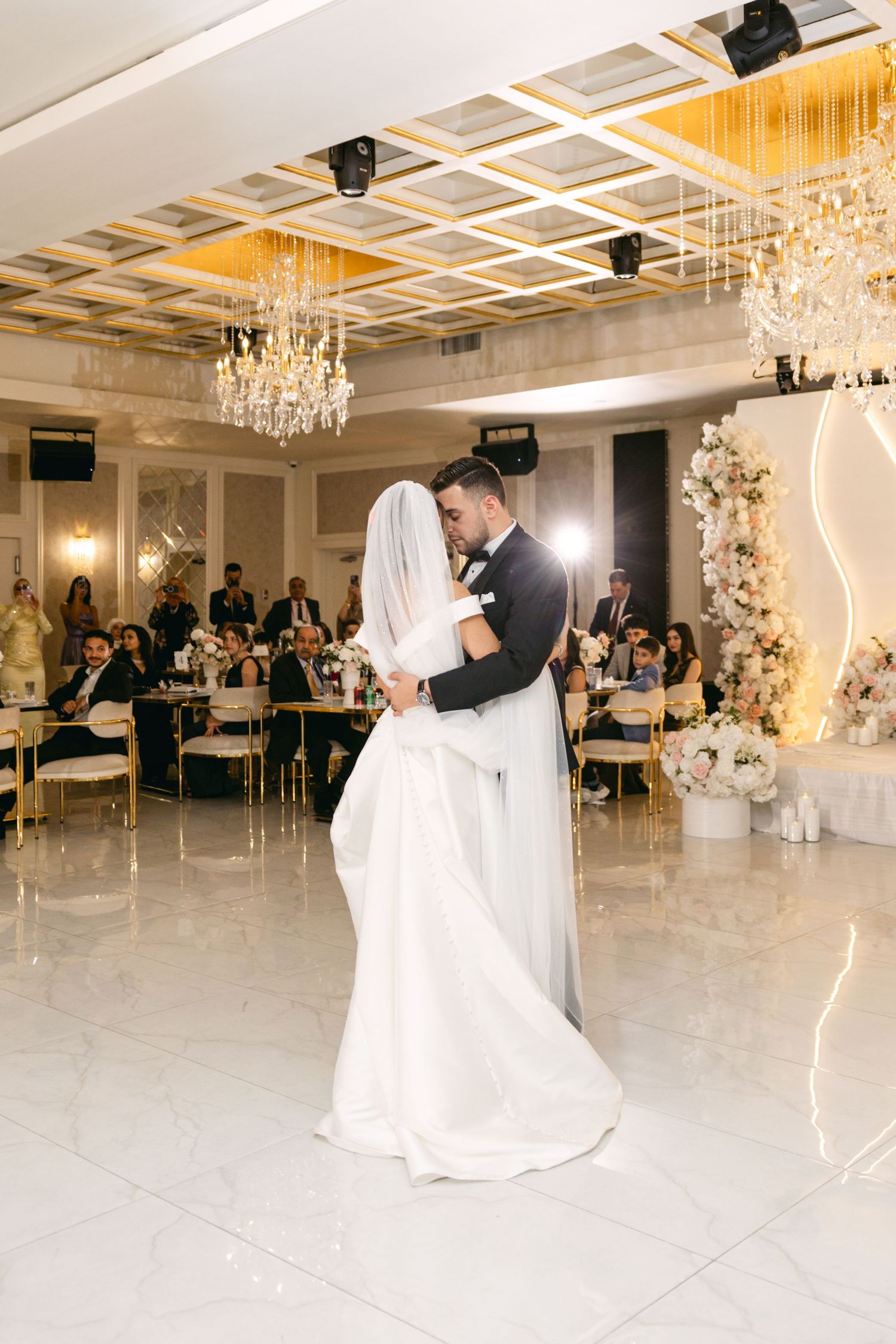 A couple shares their first dance under crystal chandeliers in an elegant white ballroom.