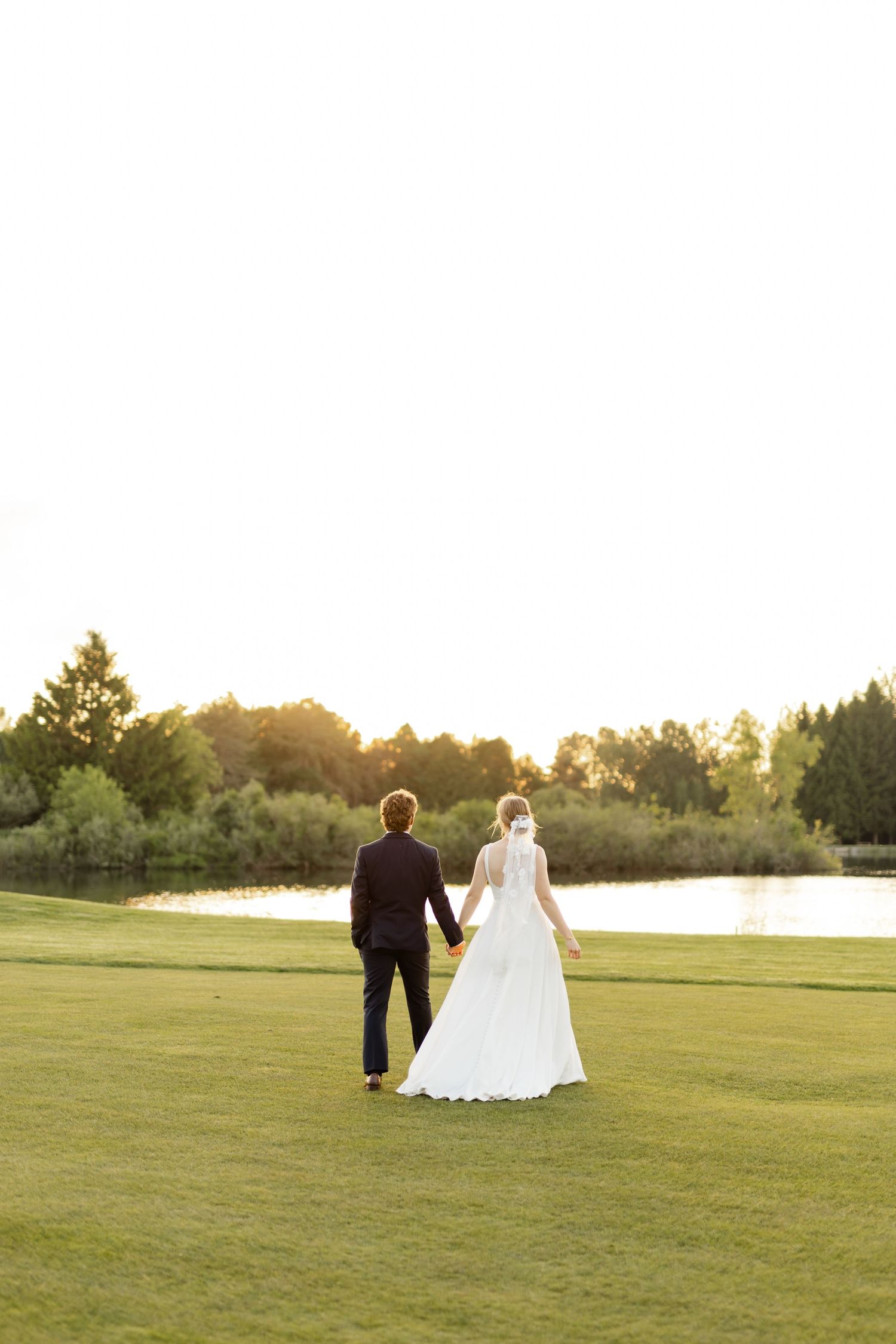 Newlyweds walk hand in hand across a sunlit green field near a lake at sunset.