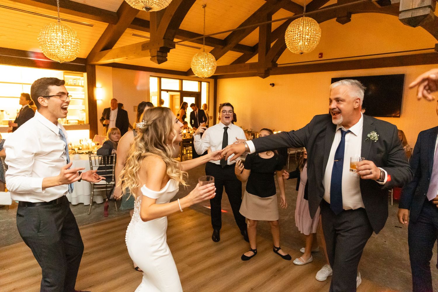 Wedding guests dancing and celebrating in a rustic venue with wooden beams and warm lighting.