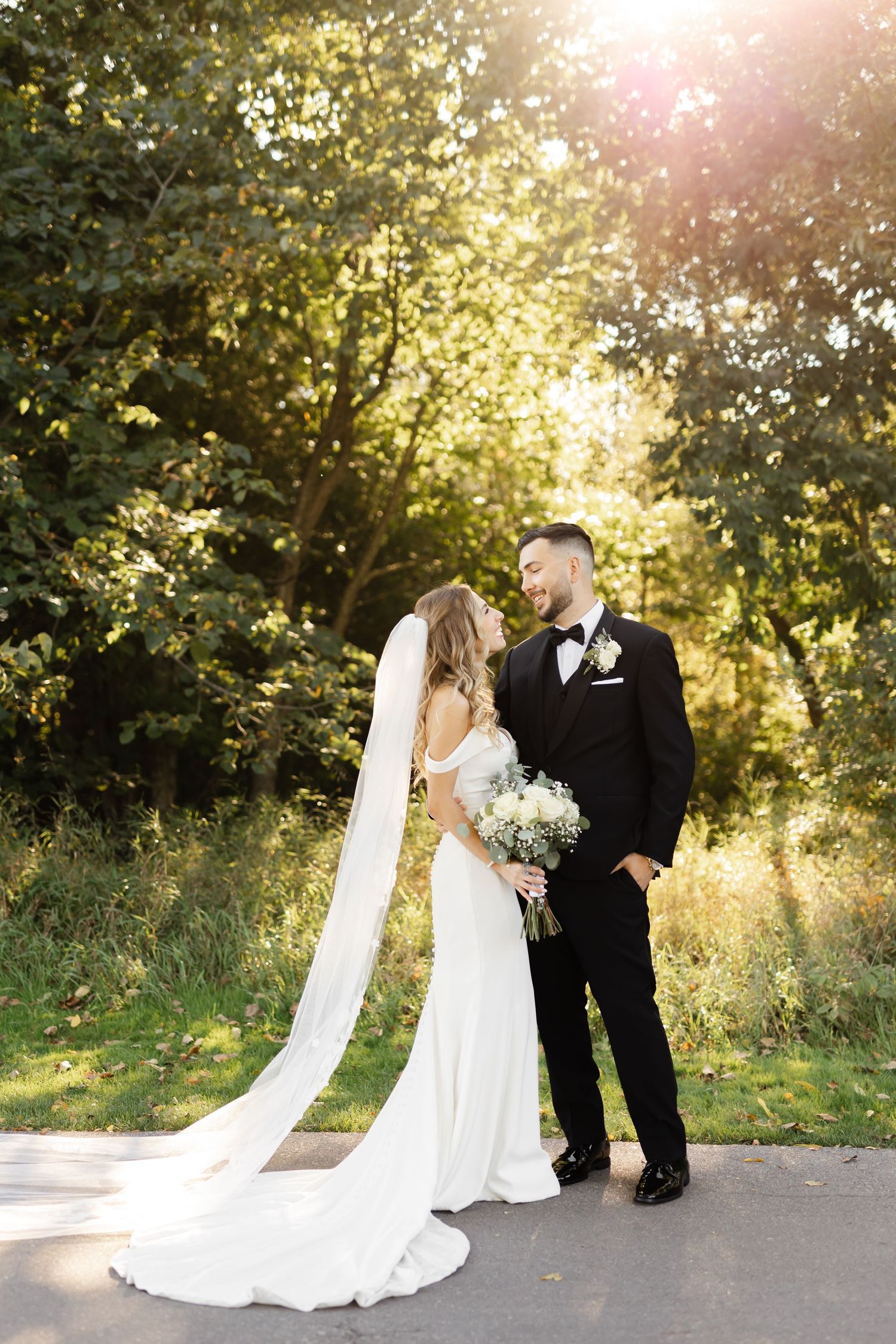 Wedding couple poses in front of sunlit trees with a long flowing white dress and black tuxedo.