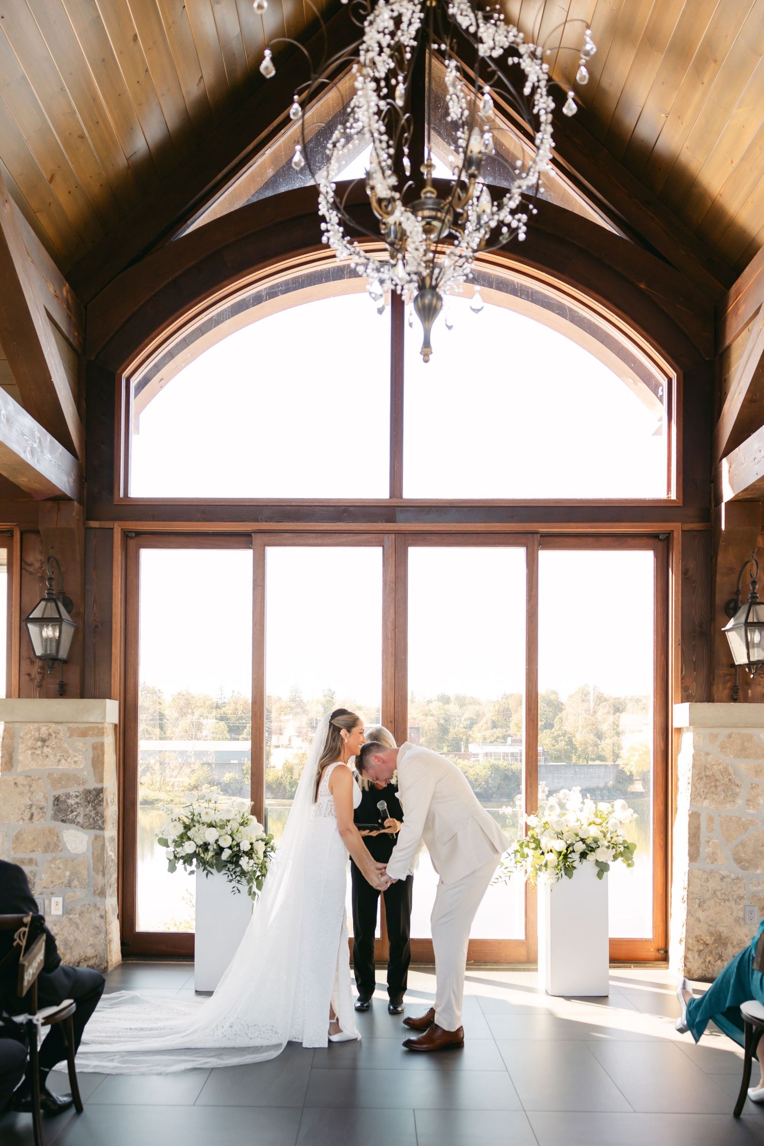 Wedding ceremony in a rustic venue with vaulted wooden ceiling and large windows overlooking scenic views.