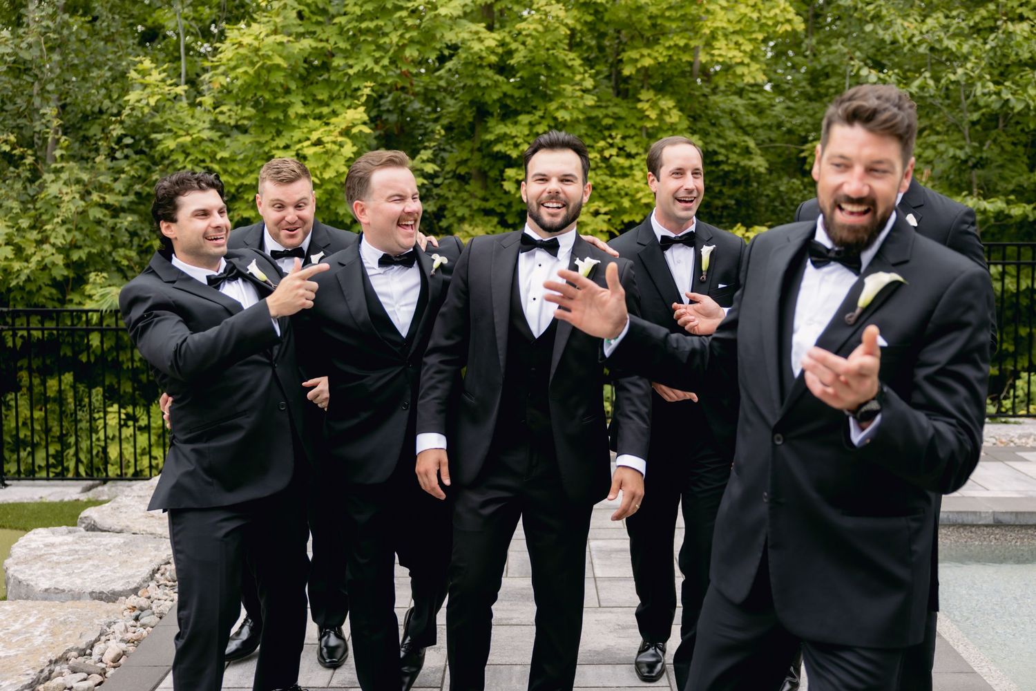 Groom and groomsmen in black tuxedos celebrating outdoors with champagne bottle near pool.