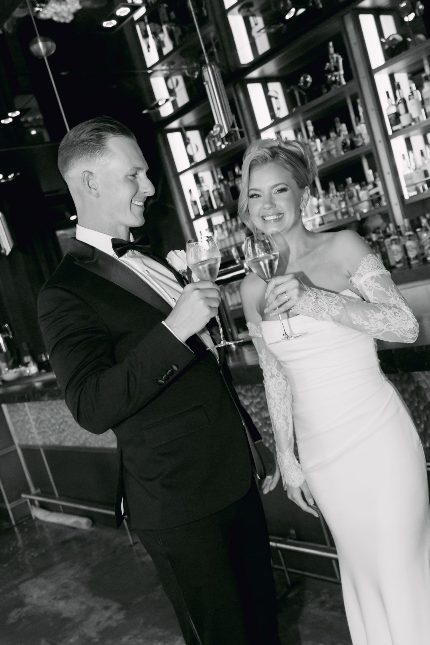 Black and white photo of bride and groom toasting champagne glasses while smiling at a bar.