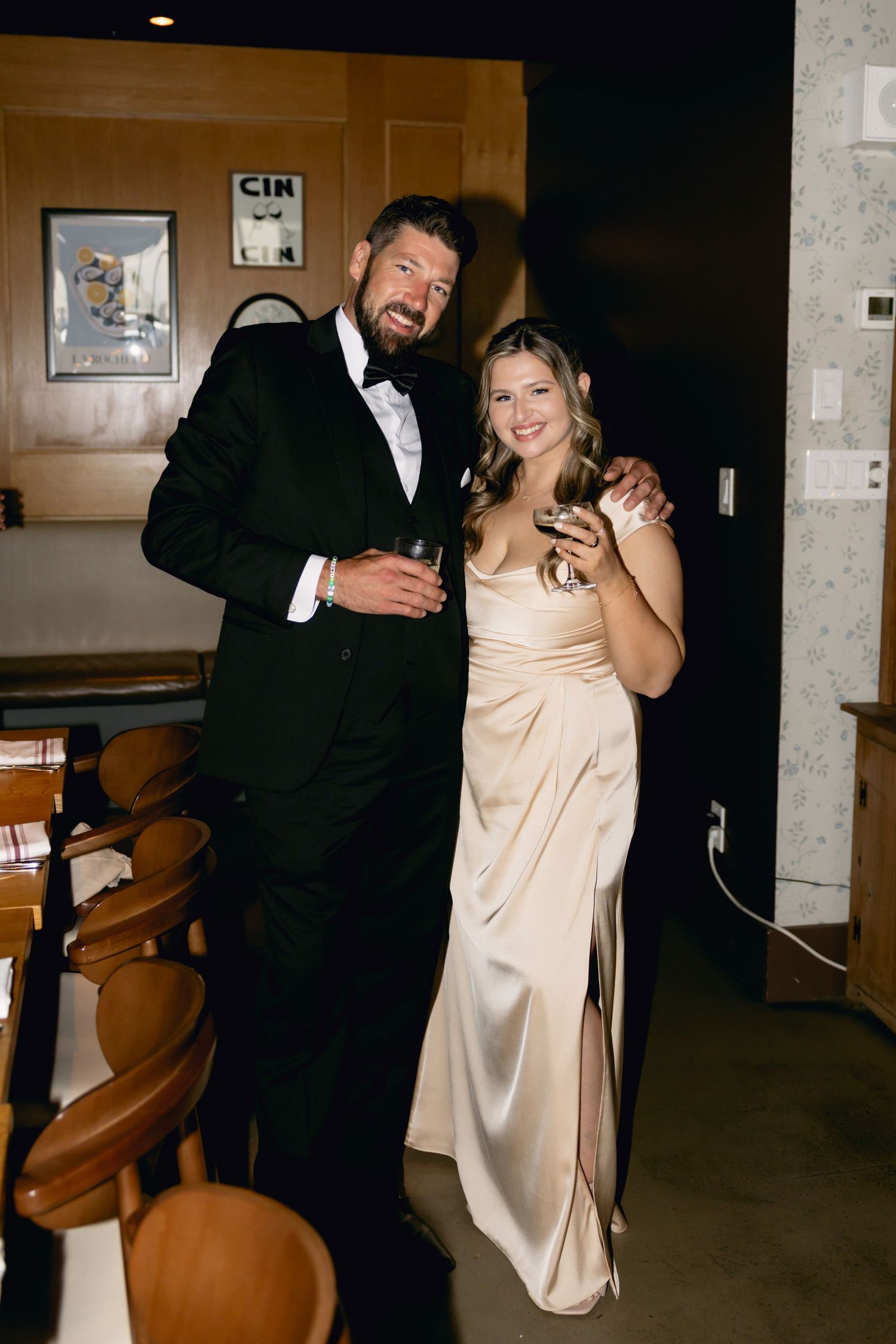 Man in black tuxedo and woman in champagne gown pose together holding drinks at reception.