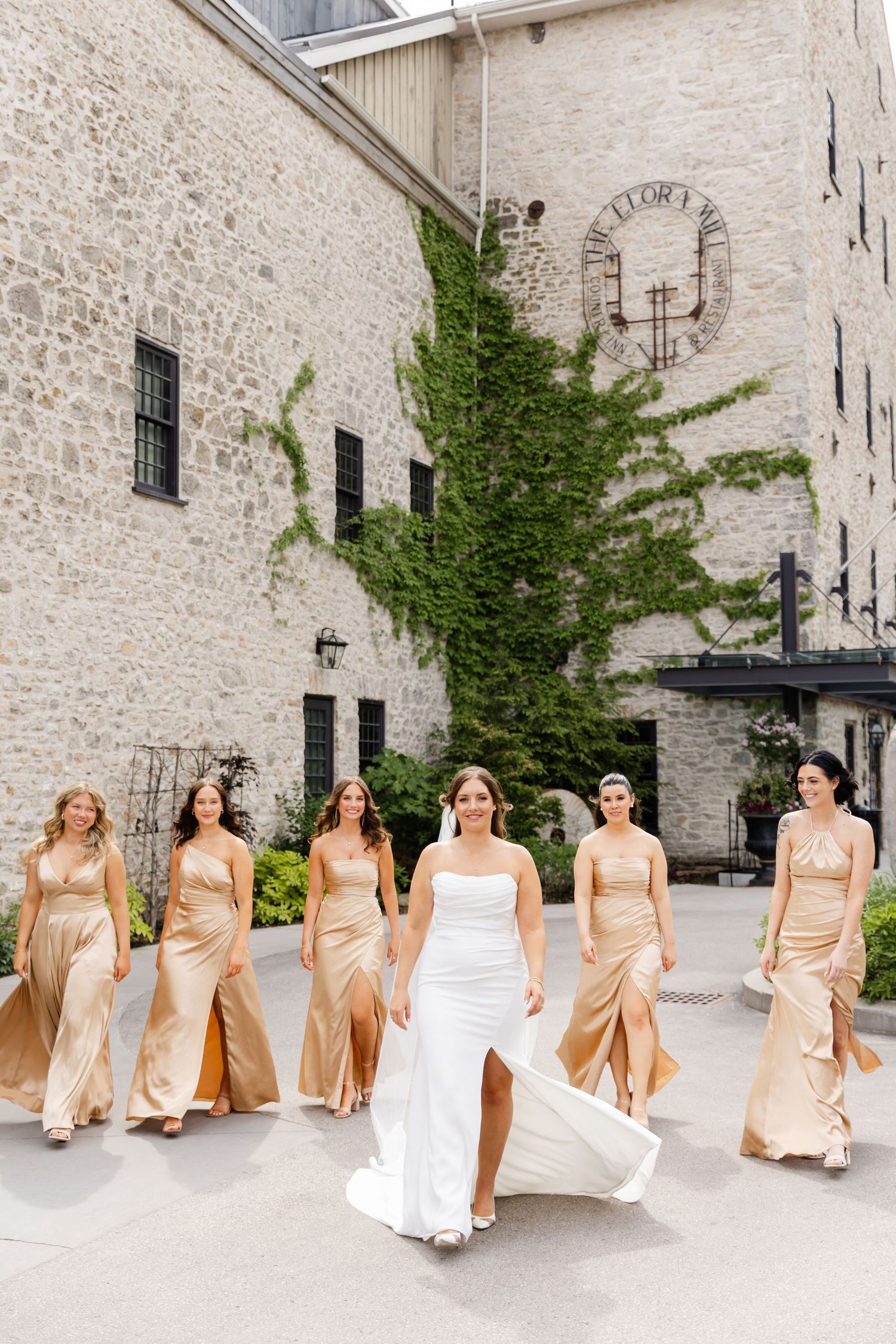 Bride in white gown leads bridesmaids in gold dresses walking confidently outside a stone venue.