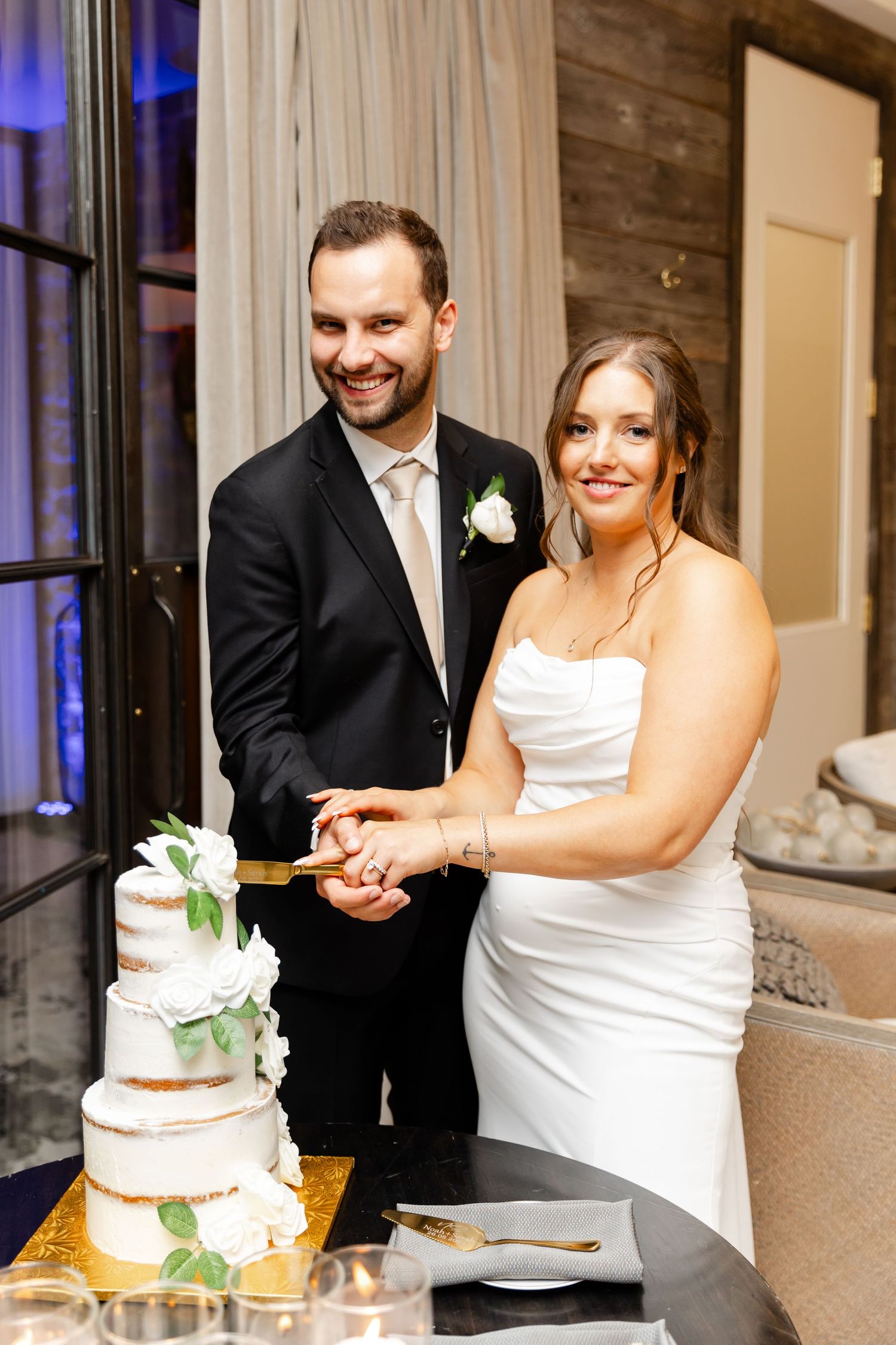 Smiling bride and groom cut their elegant three-tier white wedding cake together at the reception.