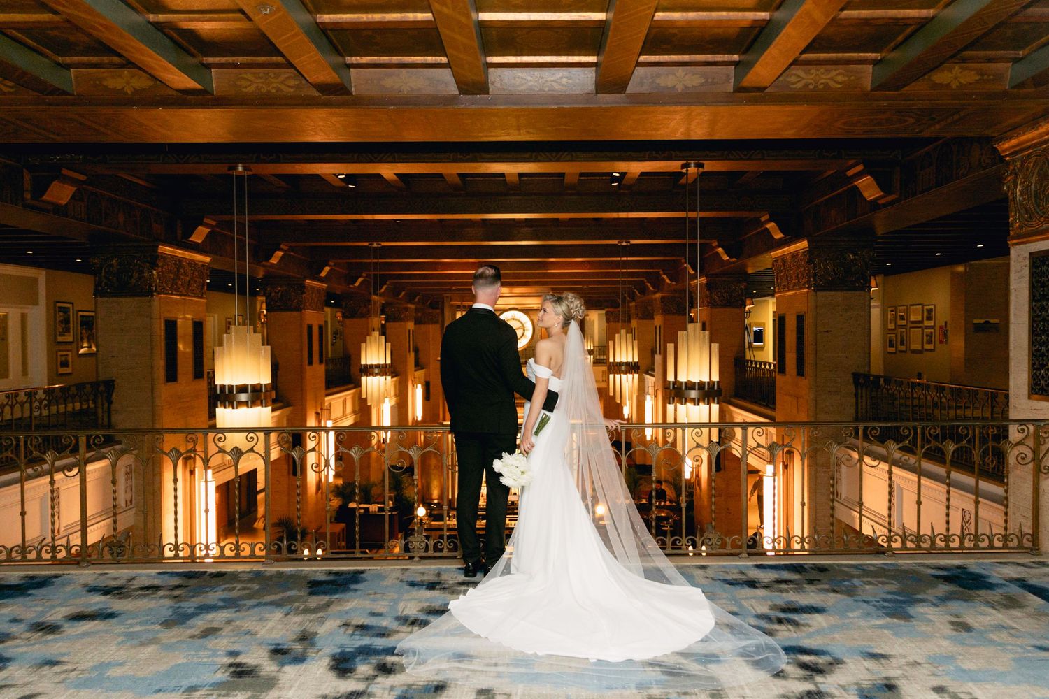 Bride and groom stand hand-in-hand in an elegant, warmly lit wedding venue with ornate architecture.