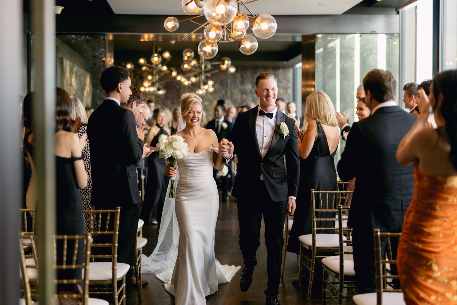 Newlyweds walk joyfully down aisle holding bouquet, guests applauding in modern chandelier-lit reception venue.