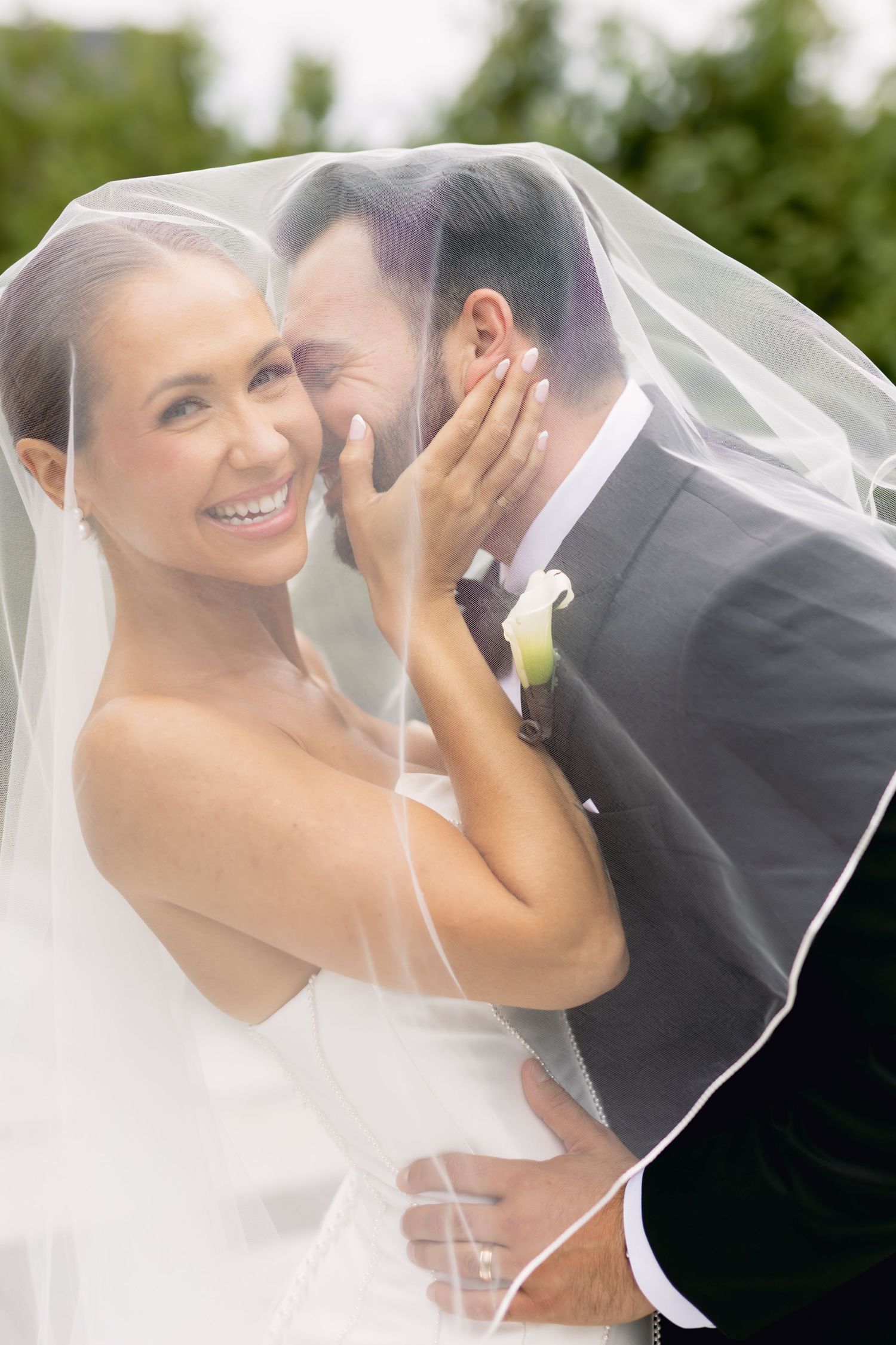 Bride smiles as groom whispers under her veil outdoors with green trees in background.