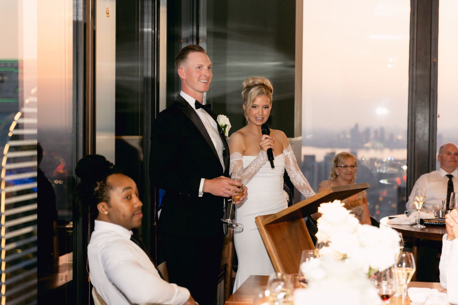 Bride and groom share a toast while giving speeches at their wedding reception with city skyline views at dusk.
