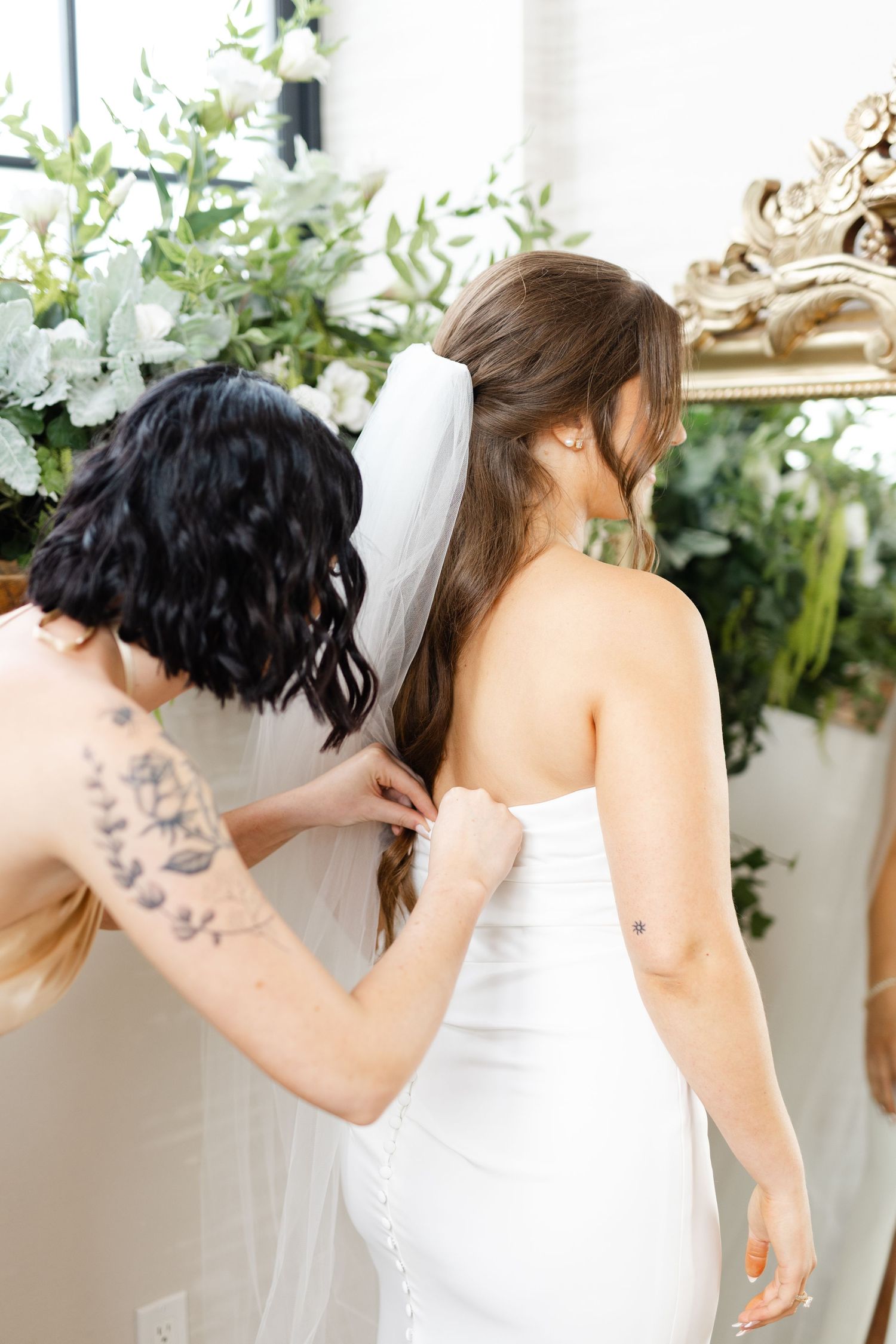 Bridesmaid with floral tattoo fastens buttons on bride's white strapless gown before wedding.
