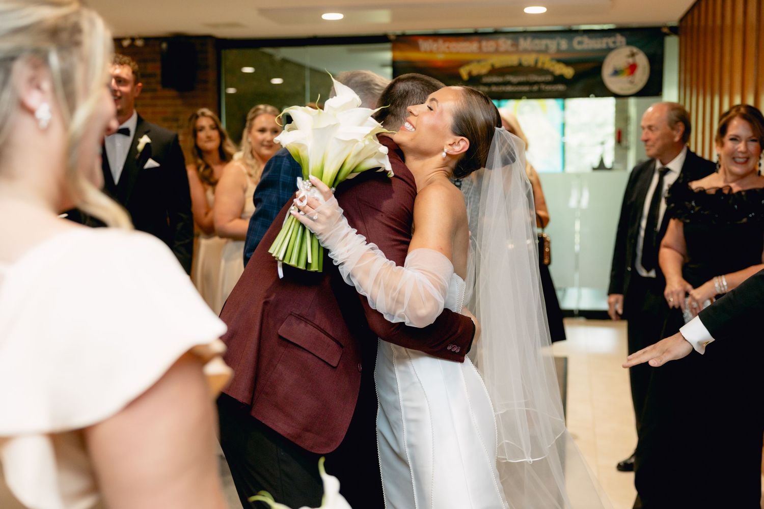 Bride embracing guest in burgundy suit while holding white floral bouquet at church entrance.