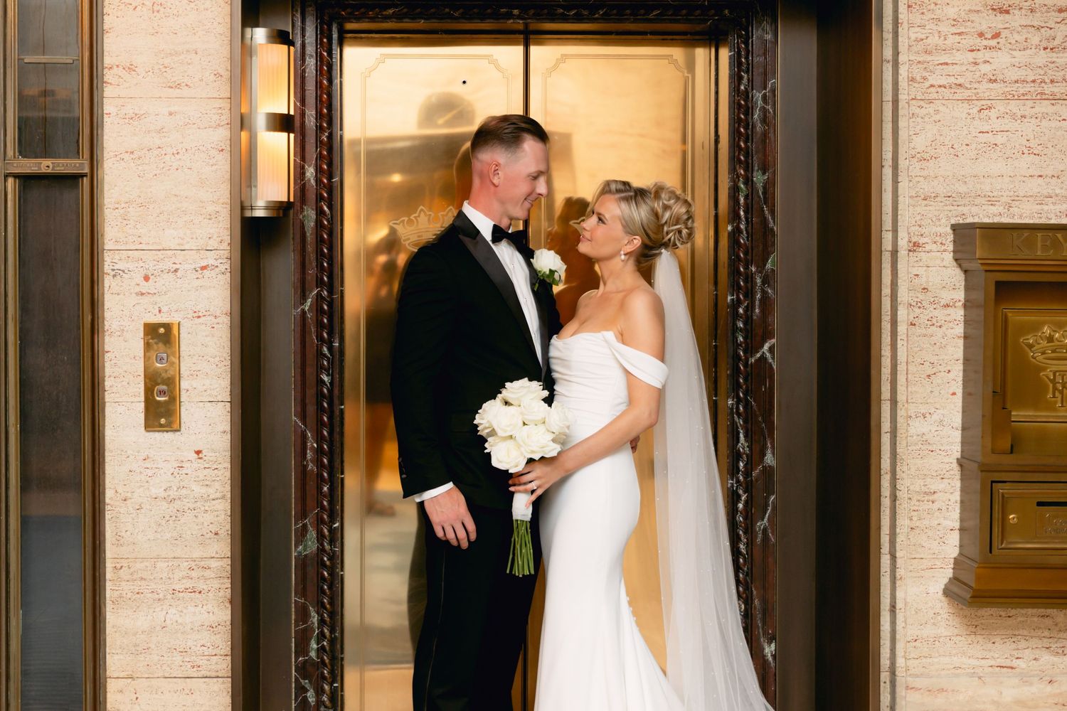 Bride and groom share a romantic moment in front of elegant gold elevator doors on their wedding day.