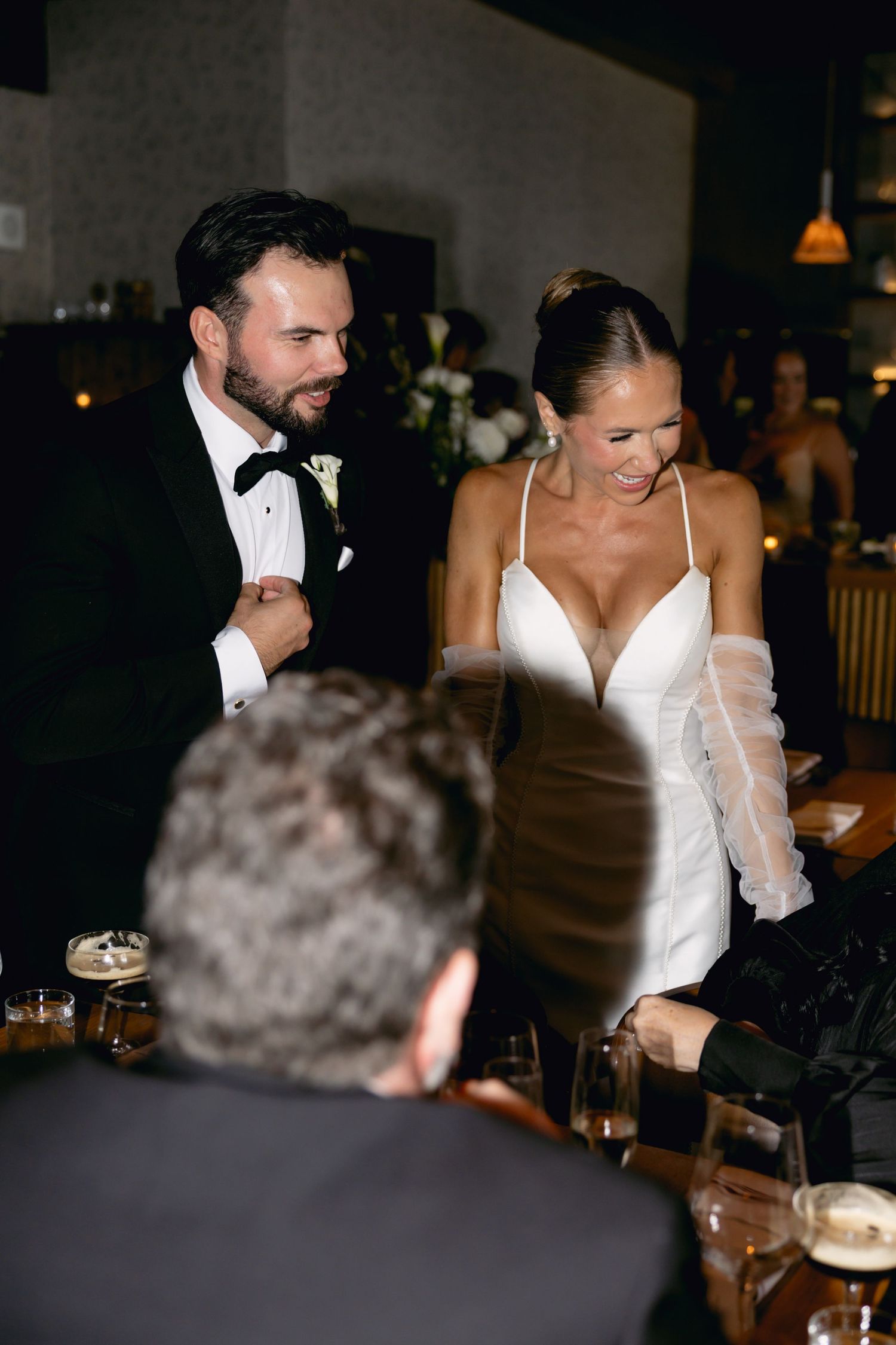 Bride in white gown and groom in tuxedo smiling at guests during wedding reception.
