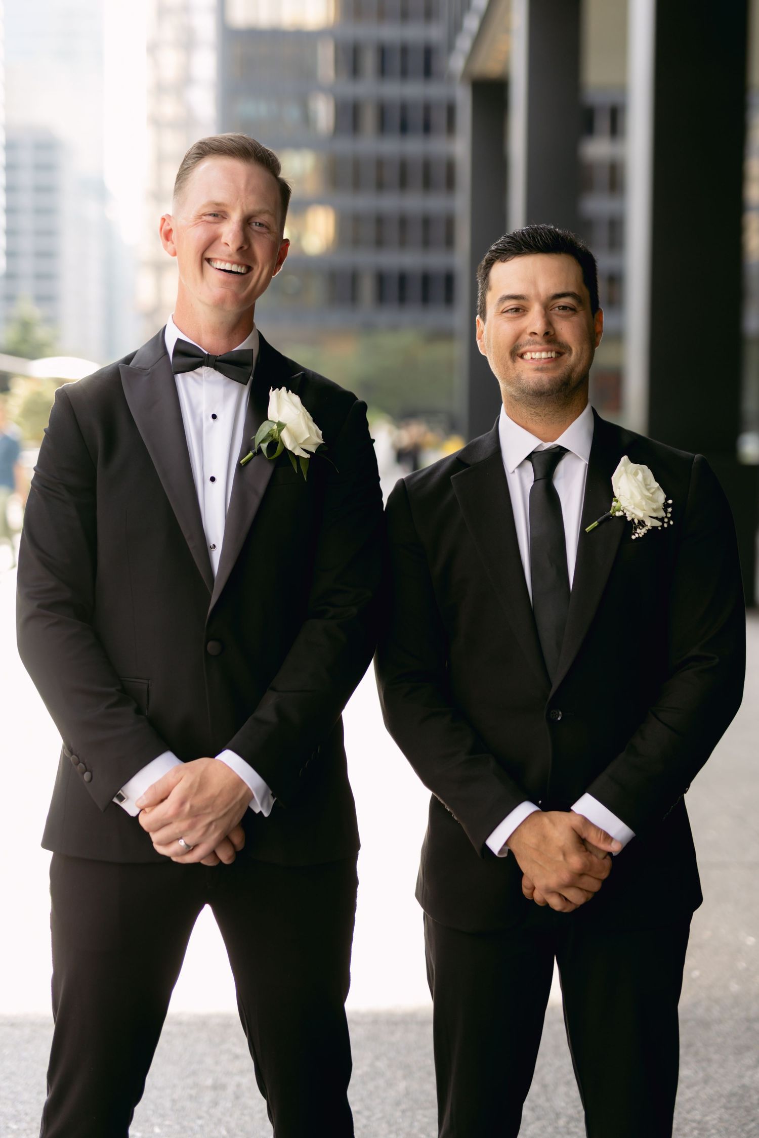 Two groomsmen in black tuxedos with white rose boutonnieres smiling outdoors in urban setting.